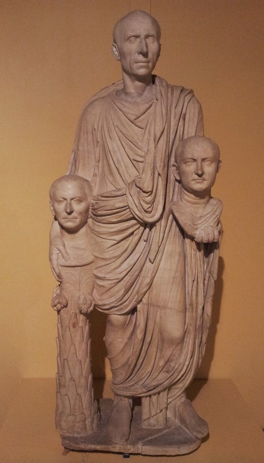 Fotografia nieznanego autora przedstawia rzeźbę nieznanego autora pod tytułem „Rzymianin trzymający popiersia swoich przodków”.  Rzeźba prezentuje łysego mężczyznę w podeszłym wieku. Jego twarz nie ukazuje emocji. Ubrany jest w długą togę sięgającą do kostek. W obu dłoniach trzyma dwa popiersia – kobiety  i mężczyzny. Fotografia rzeźby została wykonana w pomieszczeniu, które ma brązowe ściany. 