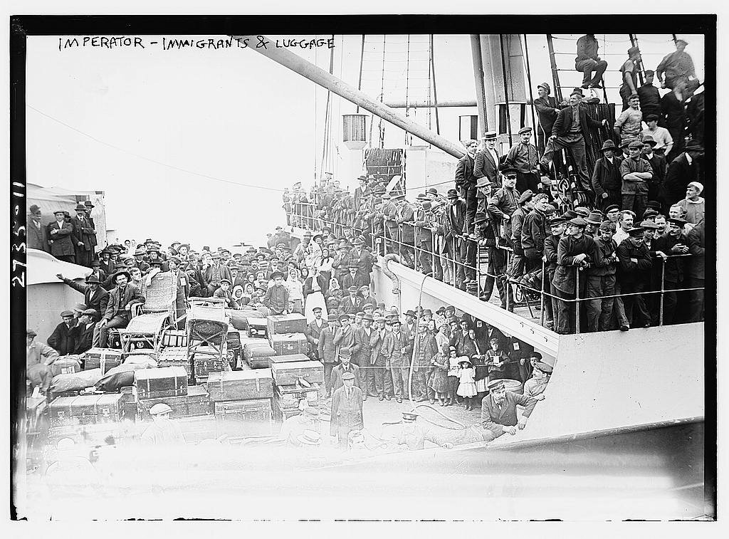 Fotografia przedstawia dwa pokłady statku pasażerskiego. Na górnym pokładzie obok barierek stoi wielu mężczyzn ubranych w robocze stroje. Na głowach mają czapki. Na dolnym pokładzie stoją mężczyźni ubrani w marynarki i spodnie. Z lewej strony ułożono wiele dużych walizek podróżnych.