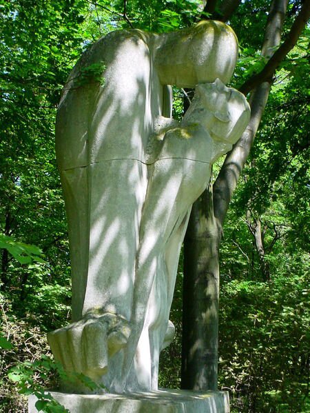 Zdjęcie przedstawia rzeźbę dwóch postaci. Stoją blisko siebie. Jedna z osób pochyla głowę do przodu. Czołem dotyka policzka osoby stojącej naprzeciwko, która odchyla głowę w tył. Rzeźba stoi na tle zielonych drzew.