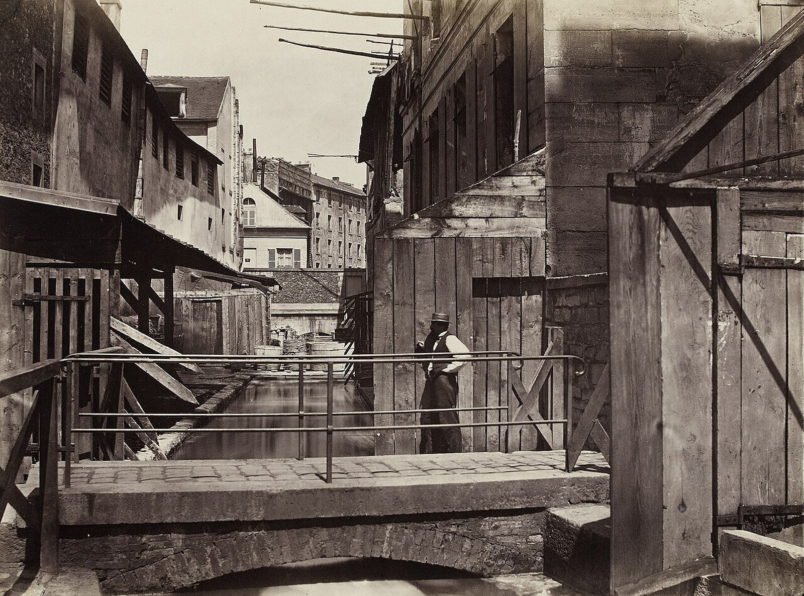 Stara czarnobiała fotografia gęstej zabudowy, po części drewnianej. Na pierwszym planie między budynkami niewielki kanał, mostek. Budynki są podniszczone.