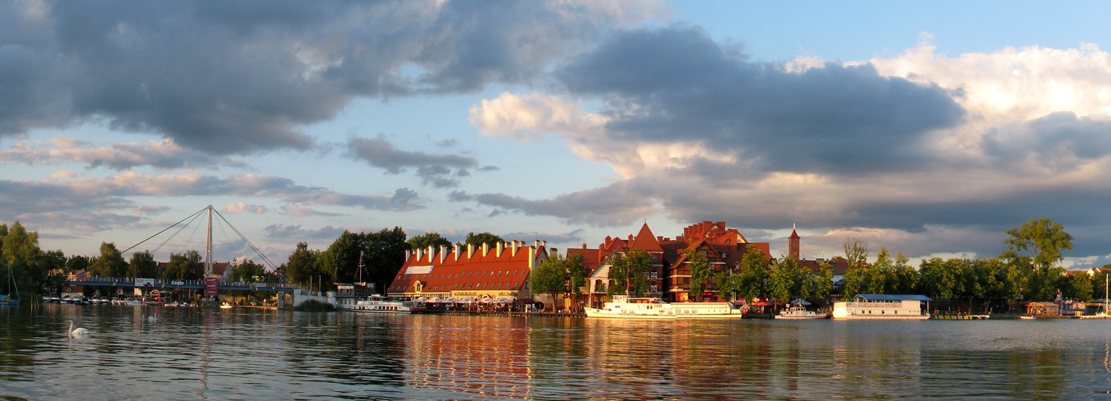 Fotografia prezentuje panoramę budynków umiejscowionych nad wodą. Na brzegu widoczne zabudowania z czerwonymi dachami oraz cumujące przy brzegu białe statki. Na dalszym planie po lewej stronie zdjęcia widać most.