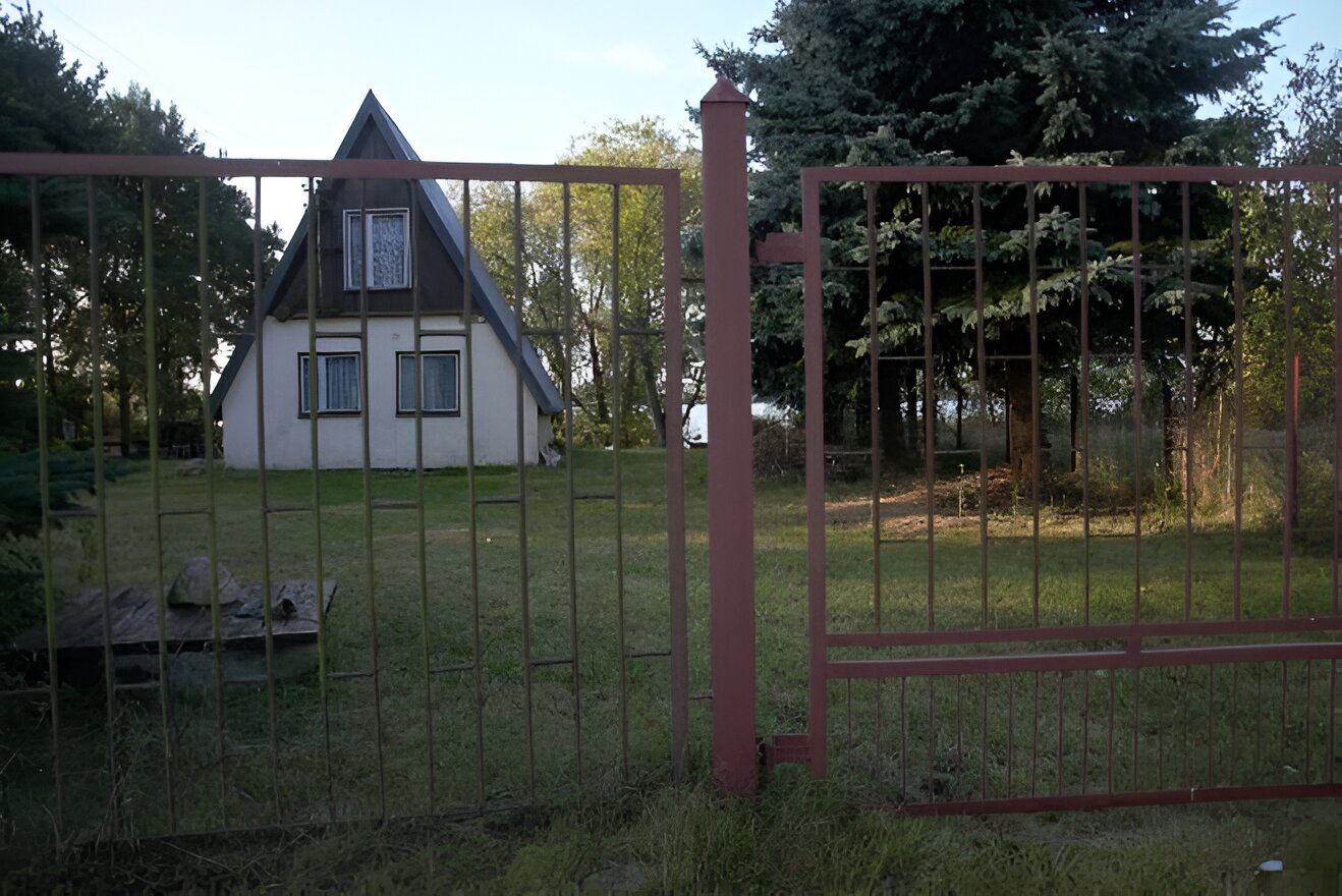 Fotografia przedstawia domek letniskowy ze spiczastym dachem, ogrodzony czerwonym ogrodzeniem, dookoła trawa i drzewa, w tle rzeka, 