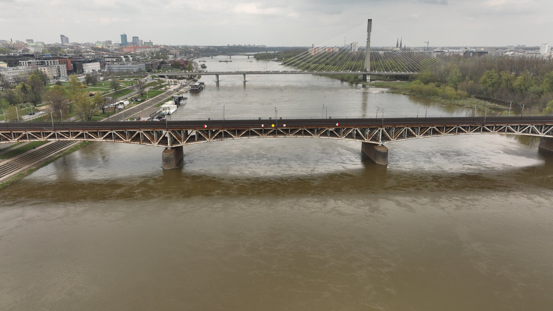  Na zdjęciu przedstawiony jest potężny most stalowy łukowy z jazdą górą, przecinający rzekę w okolicy dużego miasta. Konstrukcja mostu składa się z solidnych stalowych łuków, umieszczonych pod platformą, na której przebiegają tory kolejowe oraz znajdują się zabezpieczenia w postaci barierek. Most rozciąga się nad całym nurtem rzeki, opierając się na trzech betonowych filarach umieszczonych w wodzie. W tle widoczny jest również drugi most — stalowy łukowy, podwieszony, służący dla pieszych i samochodów. Oprócz tego na zdjęciu można zauważyć bulwary, wieżowce oraz inne budynki miejskie.