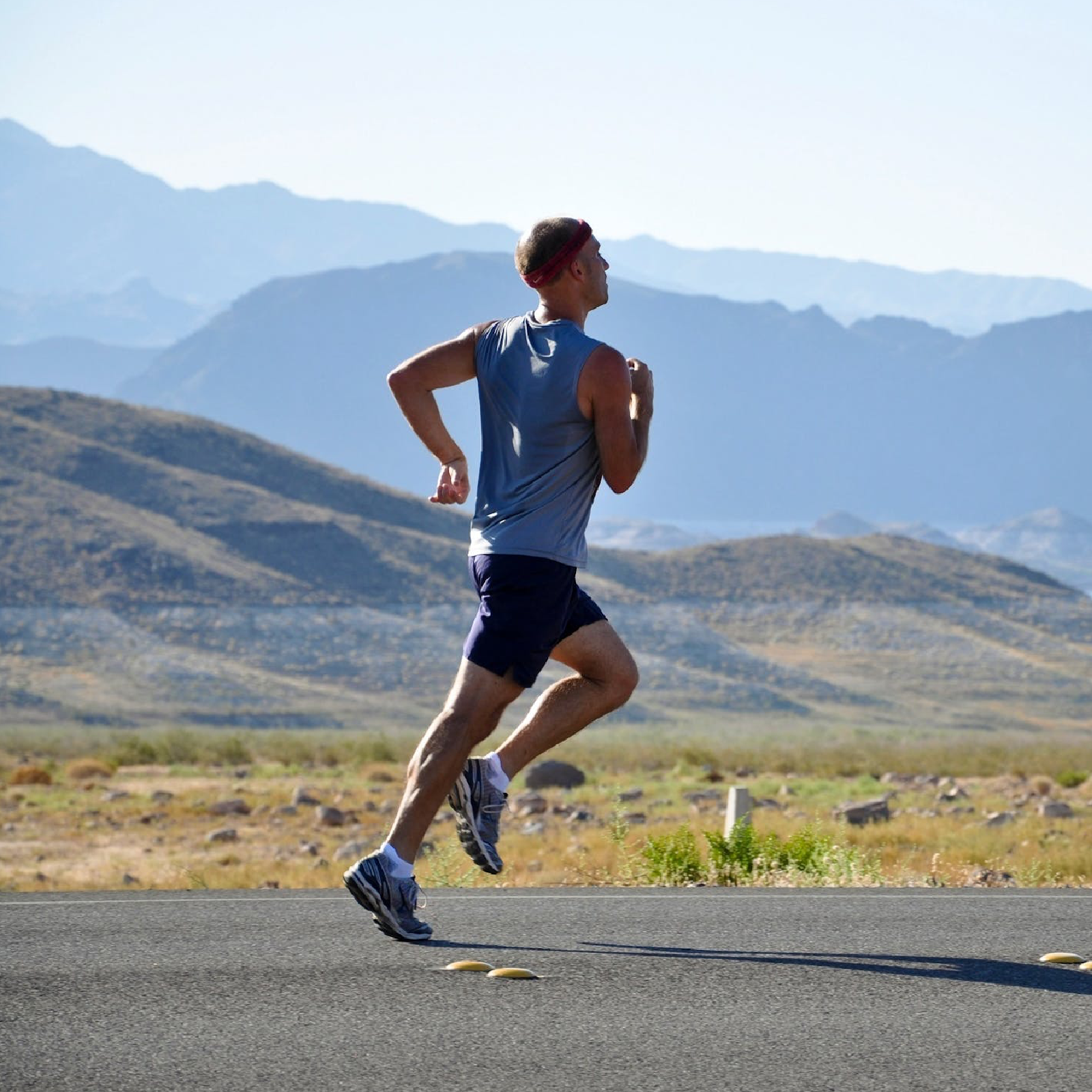 Ilustracja przedstawia mężczyznę biegnącego po asfalcie. W tle widać góry i otwarte przestrzenie. Mężczyzna ma koszulkę bez rękawków, opaskę na głowie oraz lekki strój sportowy.