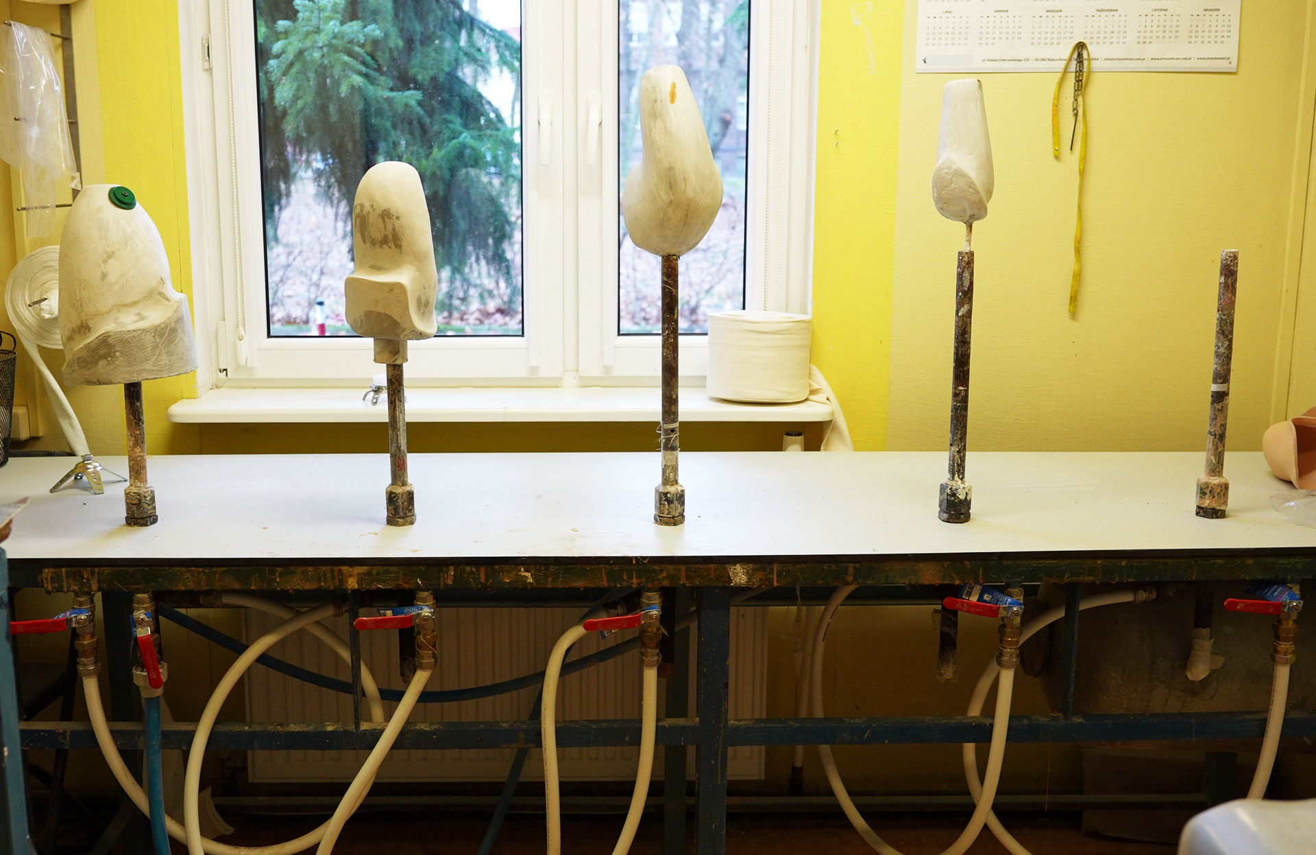 Zdjęcie przedstawia stół do laminacji. Wyposażony jest on w pięć pionowo zorientowanych rur znajdujących się w jego górnej części. Każda z rur połączona jest u dołu pod blatem w system węży i zaworów, łączących je z pompą próżniową. Na czterech rurach od lewej znajdują się pozytywy. 