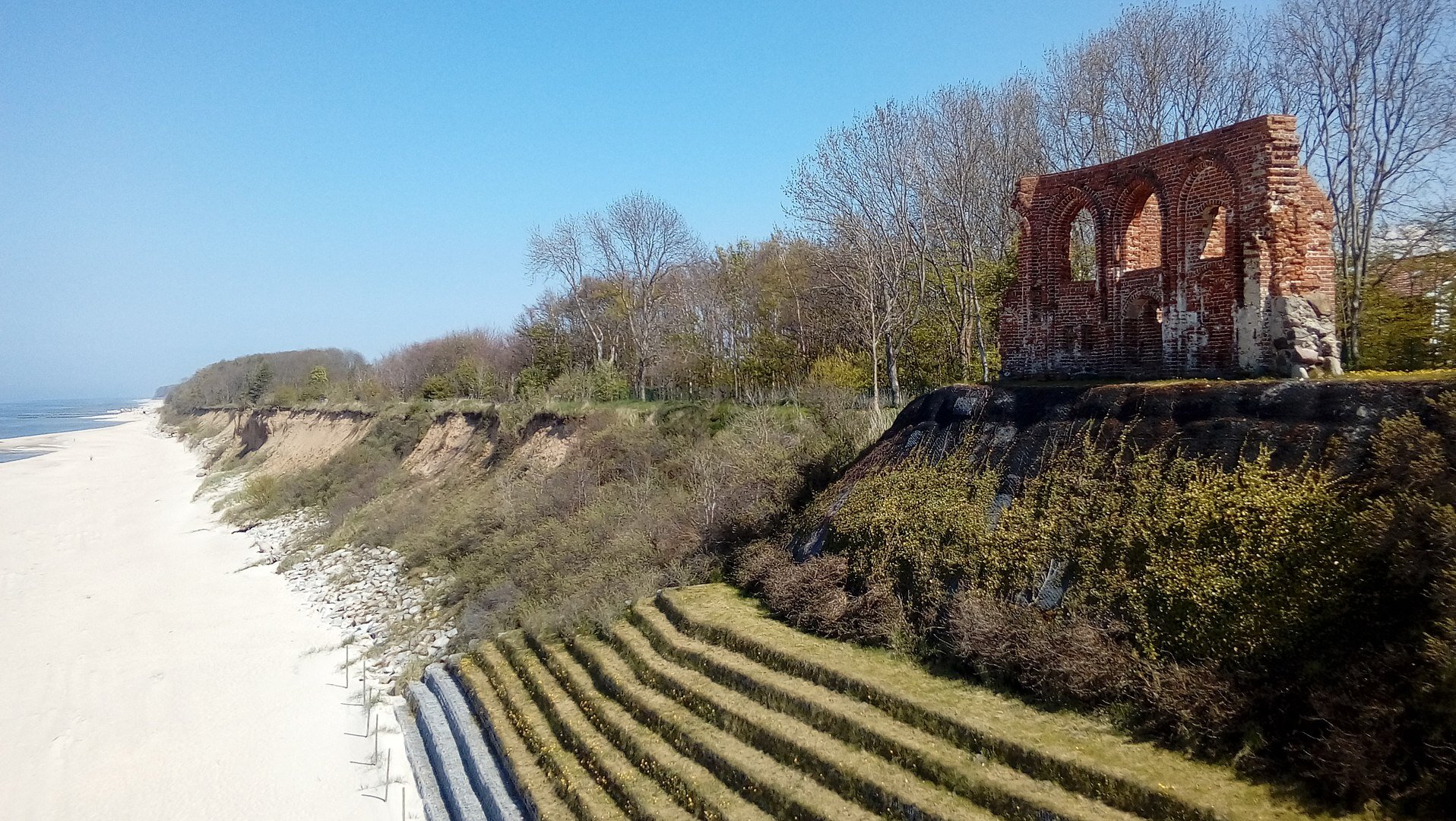 Po lewej stronie fotografii morze, dalej plaża, a za nią kilkumetrowy klif porośnięty drzewami. Na pierwszym planie, po prawej stronie, stoi na nim jedna ściana dawnego kościoła wykonana z czerwonej cegły.