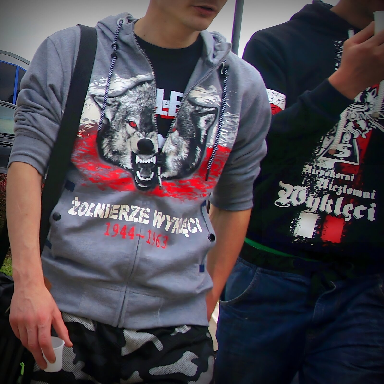 Kliknij, aby powiększyć. Na zdjęciu dwóch młodych ludzi w bluzach z motywami graficznymi nawiązującymi do żołnierzy niezłomnych. Mężczyzna po lewej stronie ma na rozpinanej szarej bluzie wizerunek głowy wilka na tle biało‑czerwonych barw i napis „Żołnierze wyklęci 1944‑1963”, mężczyzna po prawej stronie ma bluzę wkładaną przez głowę z Krzyżem Walecznych na biało‑czerwonej fladze z napisem „Niepokorni, niezłomni, wyklęci”.