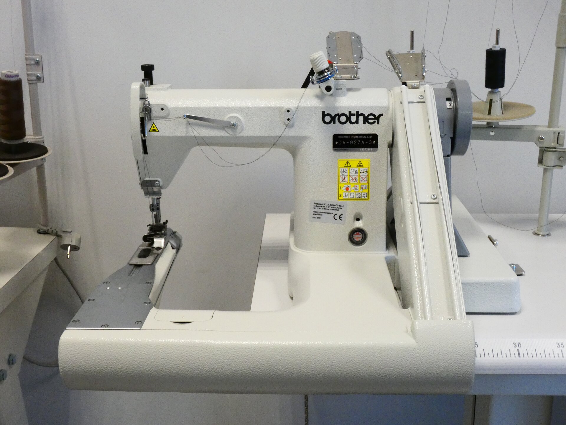 Zdjęcie przedstawia białą maszynę. Po lewej stronie znajduje się ramię z igłą. Po prawej stronie na górze maszyny nawinięte na szpule są dwie nici. 