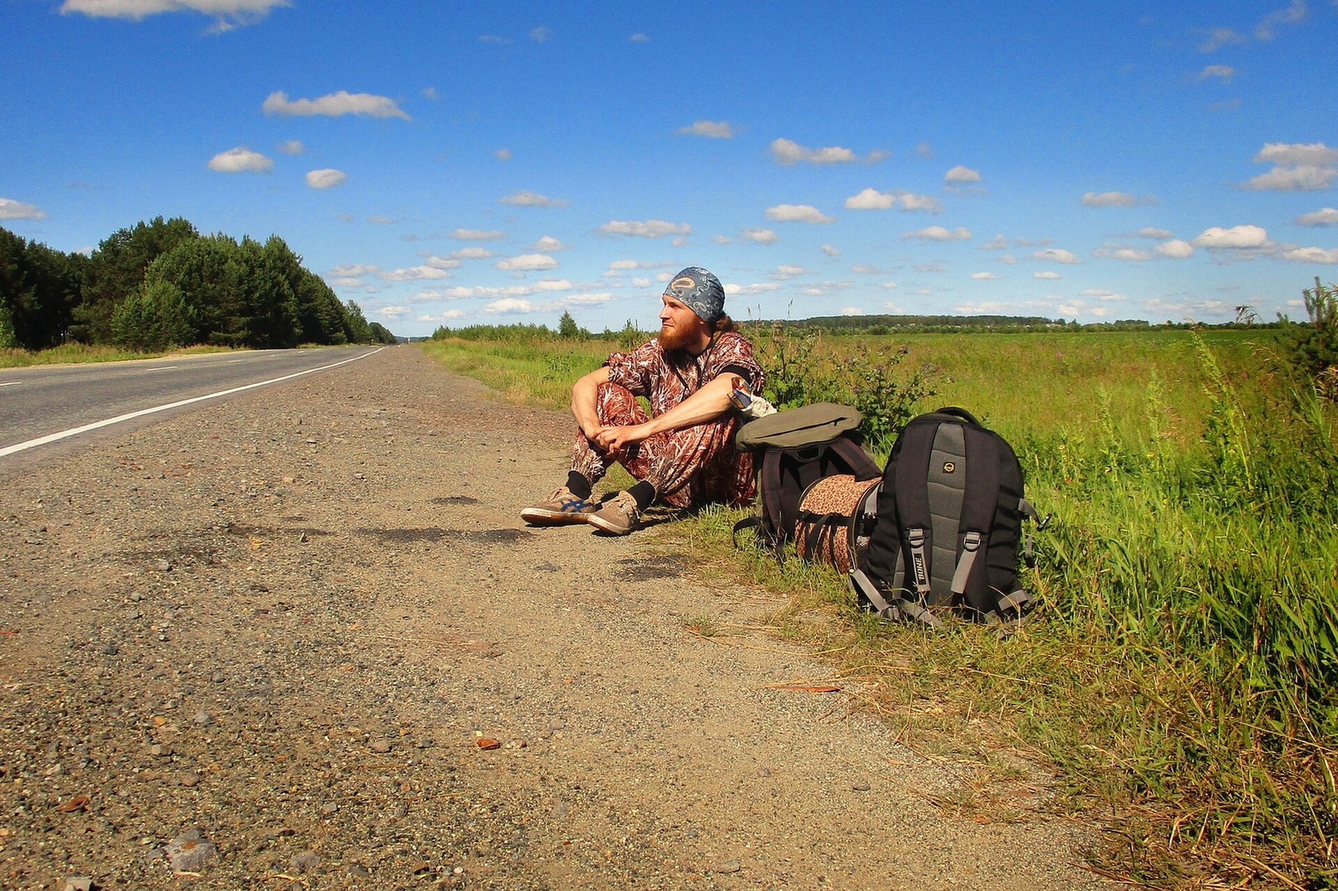 Zdjęcie przedstawia mężczyznę siedzącego na poboczu drogi biegnącej wśród łąk. Mężczyzna ma na sobie kolorowy, jednoczęściowy kombinezon, a na głowie zawiązaną chustę. Koło niego, w trawie, leżą bagaże: dwa plecaki i torba podróżna.