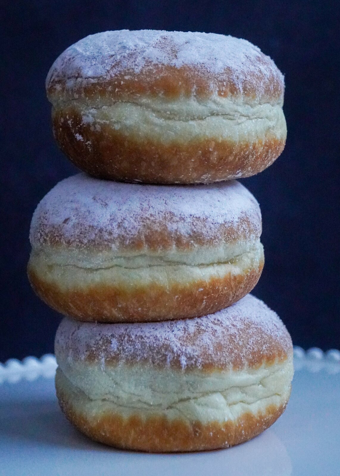 Zdjęcie przedstawia przykładowe wyroby z ciasta drożdżowego – pączki. Widoczne są trzy pączki ustawione jeden na drugim w formie wieży, leżące na białym talerzu. Pączki są posypane cukrem pudrem.