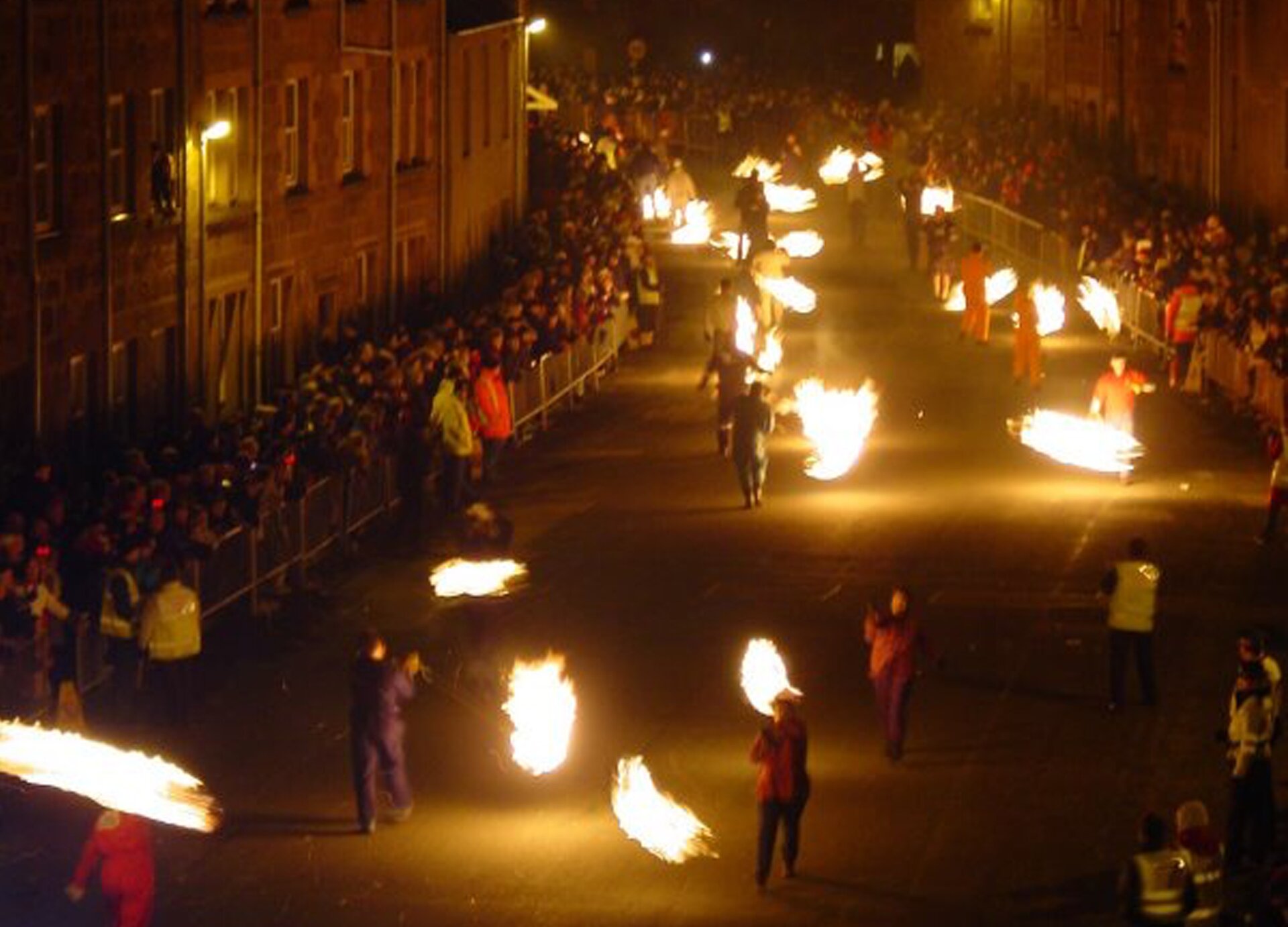 Zdjęcie przestawia pochód ulicami miasta. Uczestnicy trzymają w dłoniach płonące materiały. 