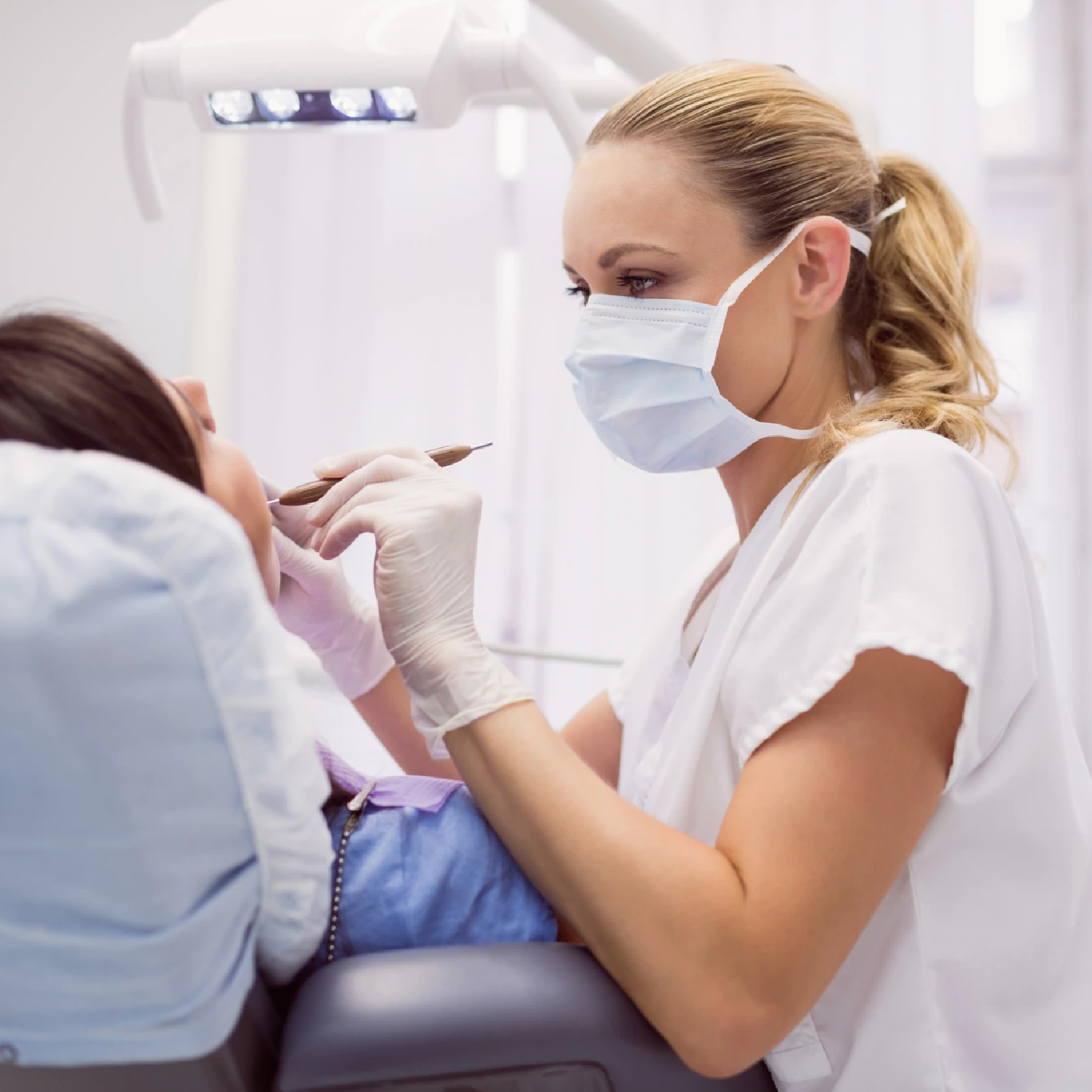 Zdjęcie przedstawia dentystkę, która obsługuje pacjenta siedzącego na fotelu dentystycznym. Dentystyka ubraną ma maseczkę, biały fartuch oraz rękawiczki jednorazowe. W dłoni trzyma dentystyczne lusterko. 