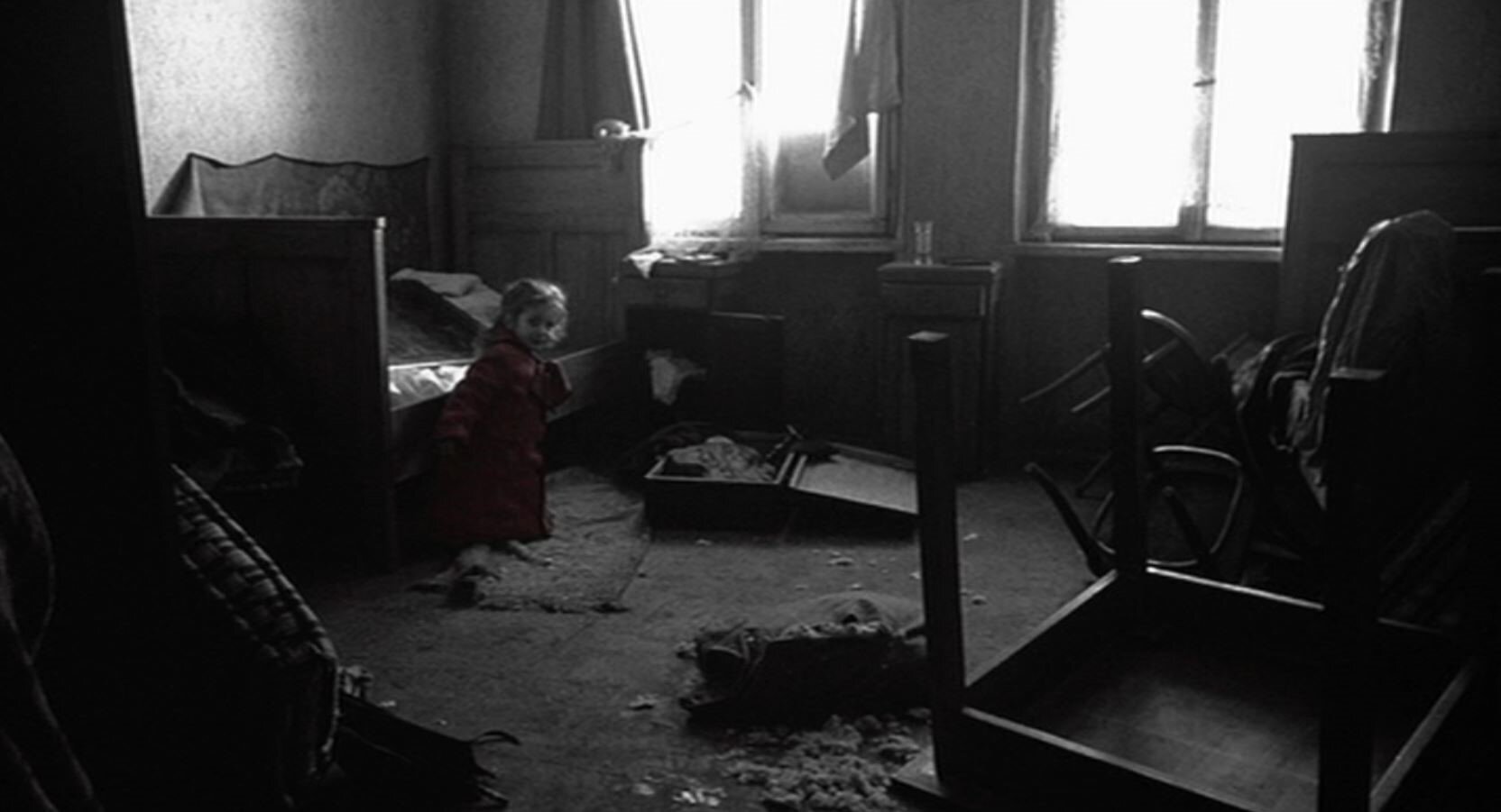 Zdjęcie czarno-białe przedstawia wnętrze pokoju. Są w nim porozrzucane rzeczy i meble. W pokoju znajduje się dziewczynka w czerwonym płaszczyku.