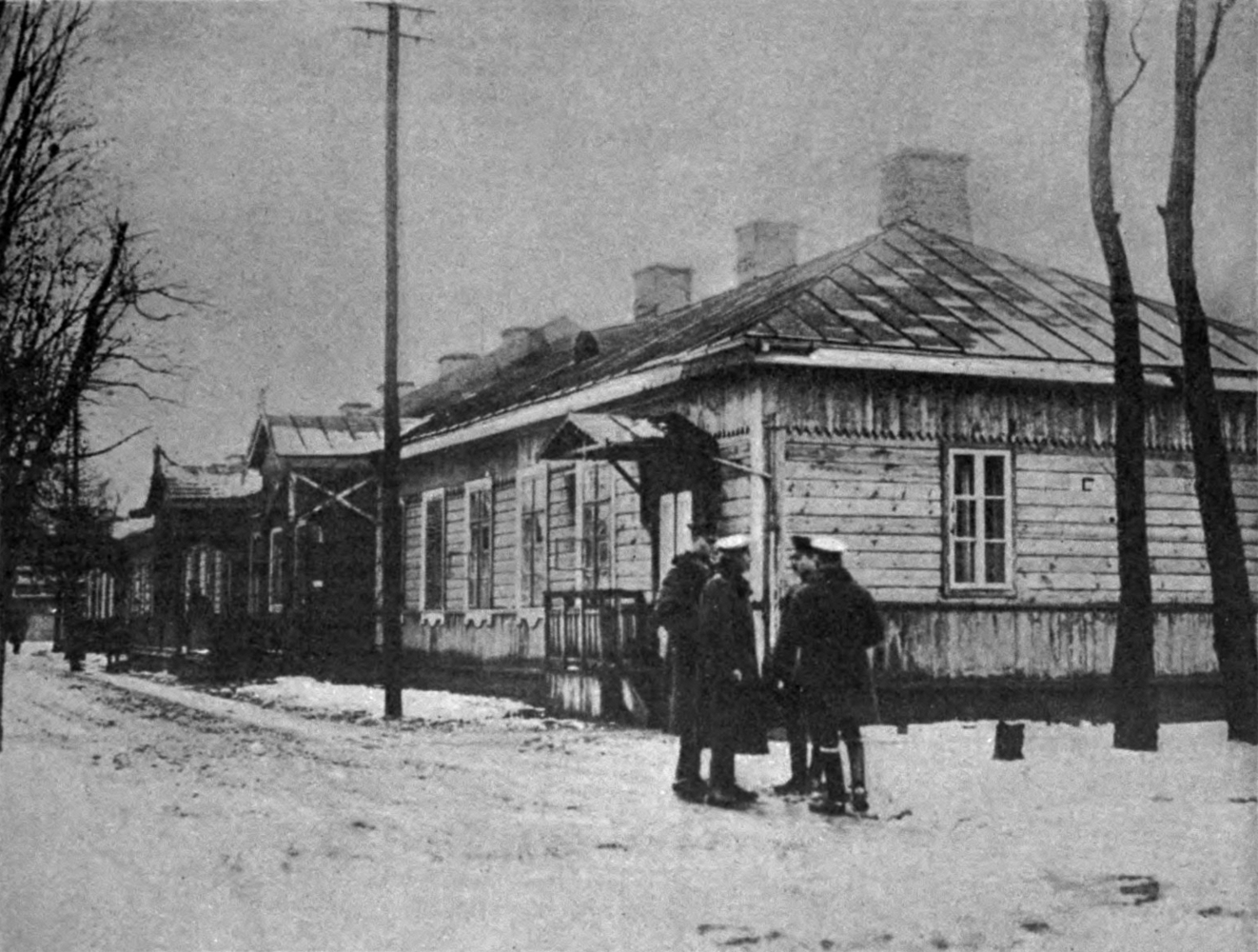 Fotografia przedstawia duży drewniany budynek z trzema kominami. Przed budynkiem czterech mężczyzn rozmawiających ze sobą. Wszędzie leży śnieg.