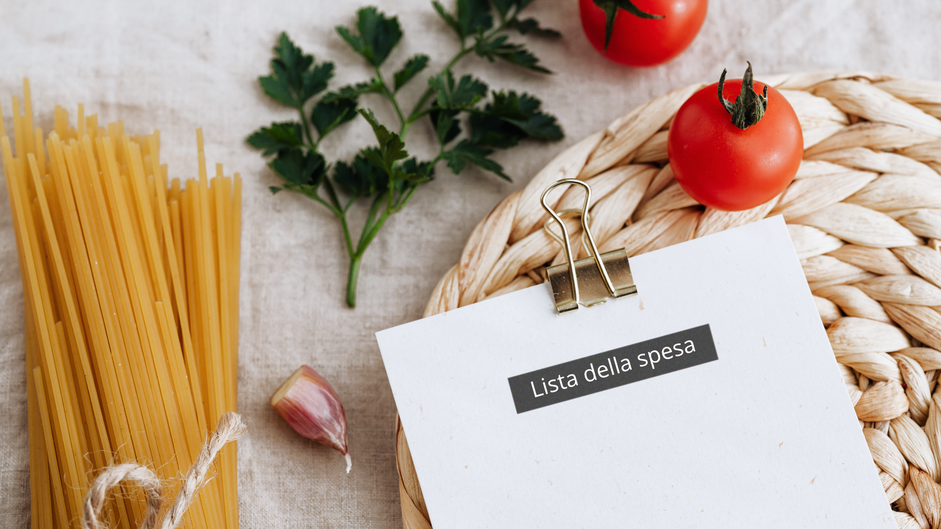 Zdjęcie przedstawia kartkę z napisem "Lista della spesa". Karta spięta jest od góry spinaczem i leży na okrągłej bambusowej podkładce. Obok kartki po lewej stronie znajdują się: suchy makaron spaghetti, ząbek czosnku. U góry znajdują się dwa pomidorki koktajlowe i gałązka pietruszki. Wszystkie te produkty wraz z kartką znajdują się na blacie stołu.