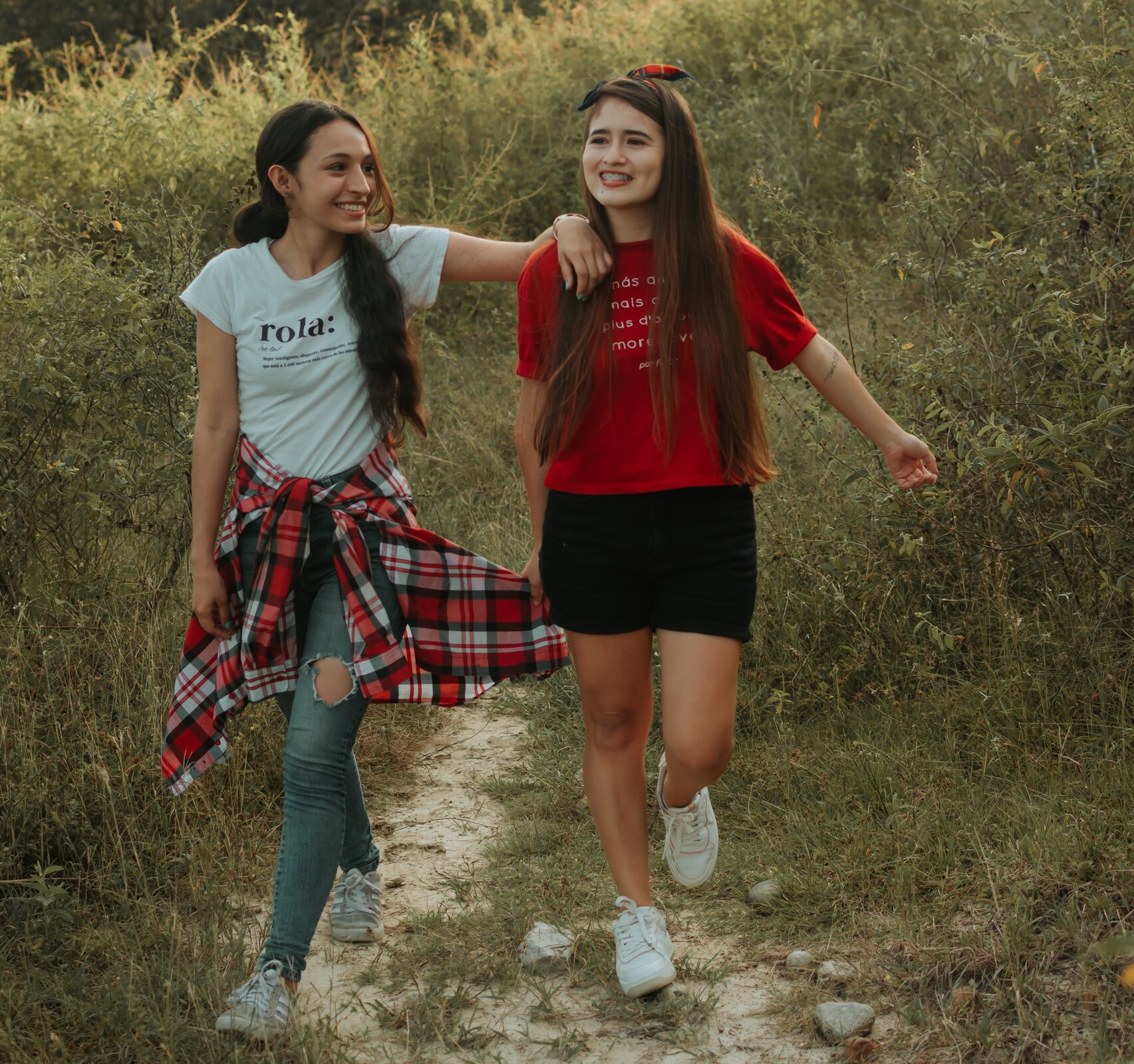 Zdjęcie przedstawia dwie dziewczyny idące ścieżką wśród wysokich traw. Jedna z dziewcząt trzyma drugiej rękę na ramieniu. Dziewczyny uśmiechają się.   