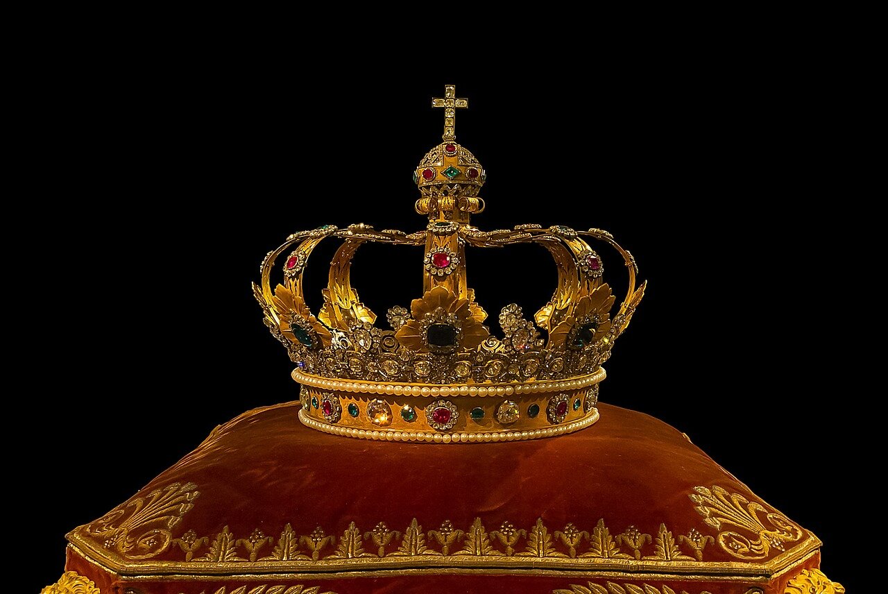 Na zdjęciu jest złota, królewska korona bogato inkrustowana kamieniami szlachetnymi. Korona leży na ozdobnej, welwetowej poduszce. 