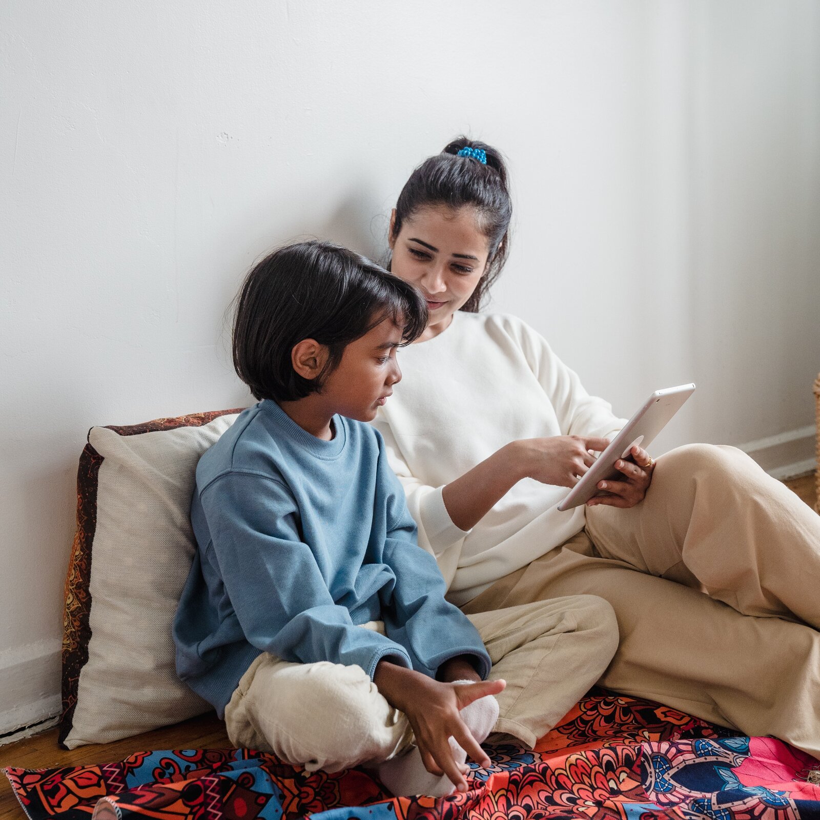 Zdjęcie przedstawia kobietę i chłopca w wieku około 9 lat. Oboje siedzą na podłodze pod ścianą. Kobieta patrzy w kierunku chłopca a w rękach trzyma tablet pokazując coś na ekranie.