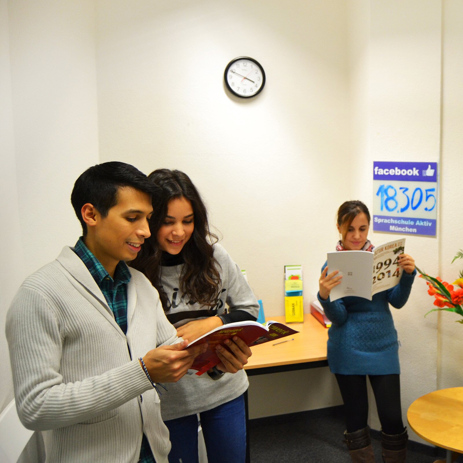 Zdjęcie przedstawia trzy młode osoby stojące w pomieszczeniu. Od lewej widoczny jest chłopak z krótkimi, ciemnymi włosami, ubrany w koszulę w kratę i rozpinany szary sweter, który w lekko ugiętych dłoniach trzyma otwartą książkę. Obok niego stoi dziewczyna z długimi, ciemnymi włosami, ubrana w szarą bluzę i dżinsy, która spogląda na strony otwartej książki trzymanej przez chłopaka. Po prawej stronie w głębi widoczna jest dziewczyna z ciemnymi, związanymi włosami, ubrana w niebieski sweter i czarne legginsy, która w dłoniach trzyma otwartą książkę. Ma lekko pochyloną głowę i wzrok utkwiony na otwartych stronach. W tle widoczne jest biurko, zegar, stolik z kwiatami.