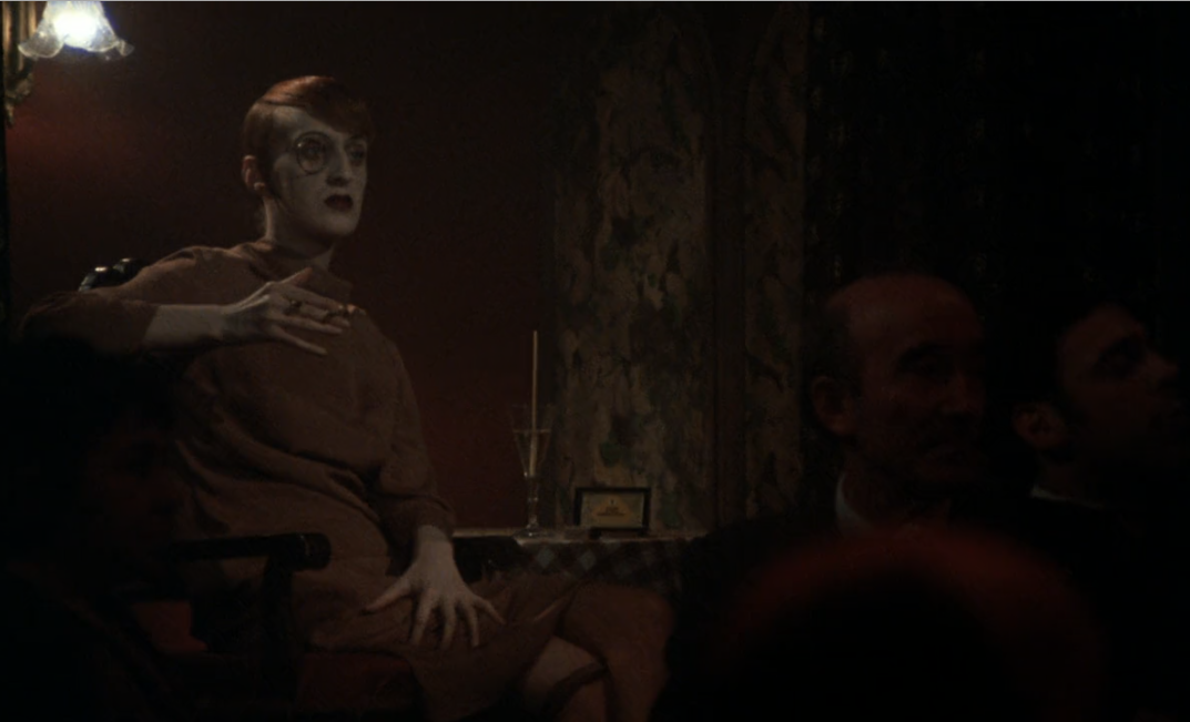 Zdjęcie przedstawia postać pomalowaną na biało w brązowej sukni. Siedzi przy stole, ma okrągłe okulary i pali papierosa.