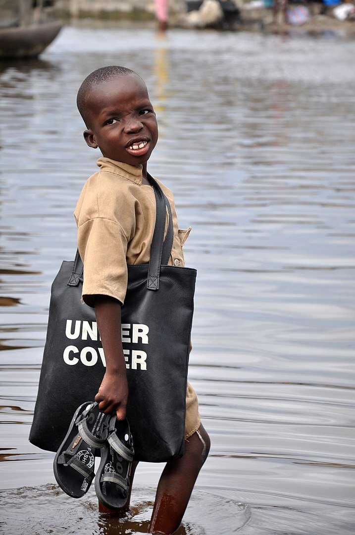 Zdjęcie przedstawia czarnoskóre dziecko, które stoi w wodzie po kostki. W rękach trzyma sandały. Na ramieniu ma zawieszoną torbę.