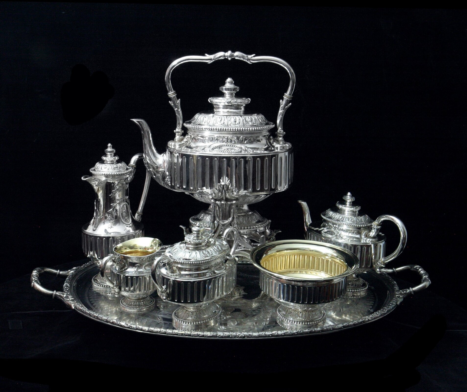 Na zdjęciu widać różne elementy srebrnej zastawy kawowej ułożonej na tacy. W centralnej części znajduje się dzbanek kawowy na stopce, obok niego mlecznik i dzbanek na herbatę, przed dzbankiem cukiernica, mlecznik i misa.