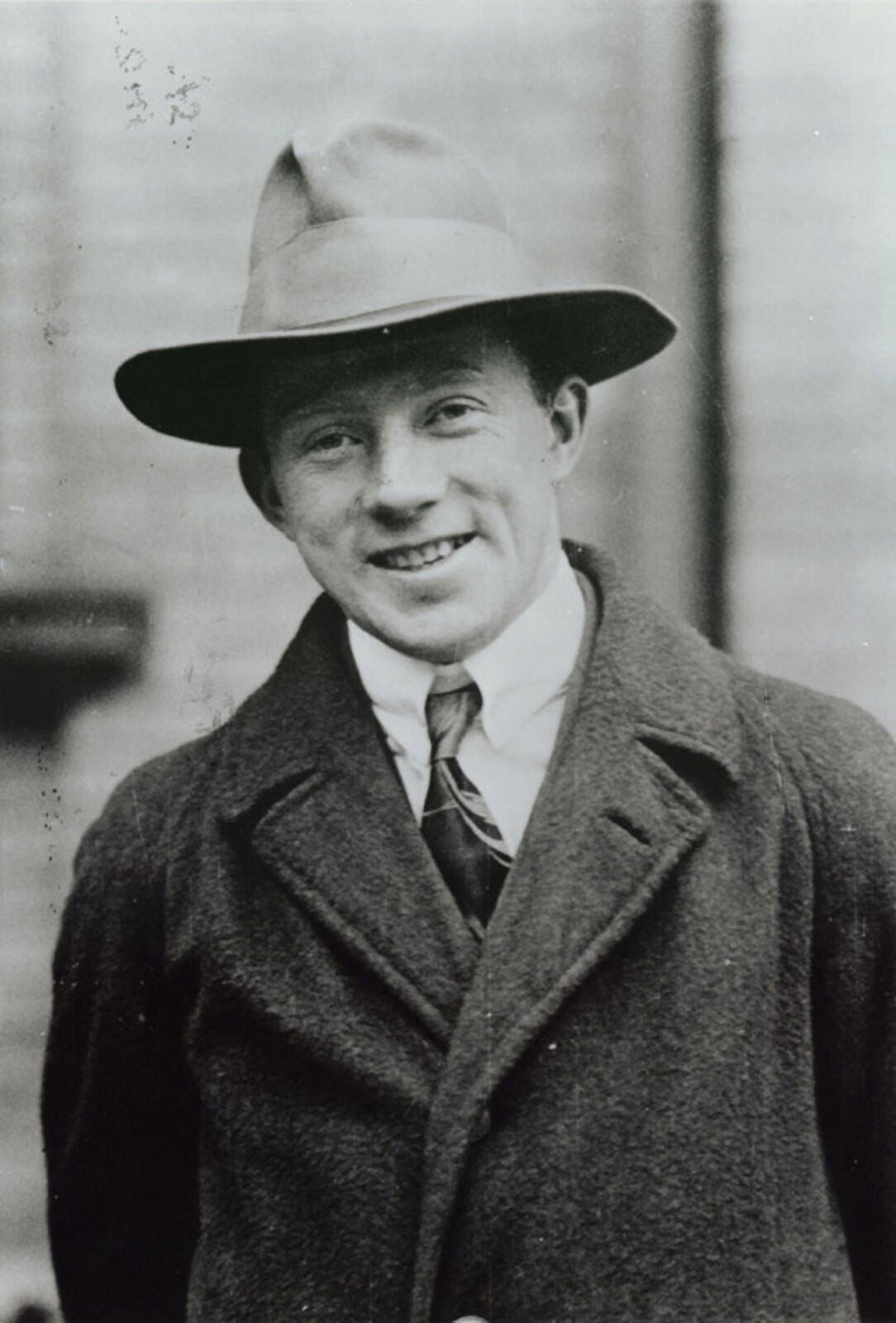 Rys. 6. Czarno‑białe zdjęcie poglądowe przedstawia Wernera Heisenberga. To młody, szeroko uśmiechnięty mężczyzna, w płaszczu, marynarce, białej koszuli, krawacie i w kapeluszu.