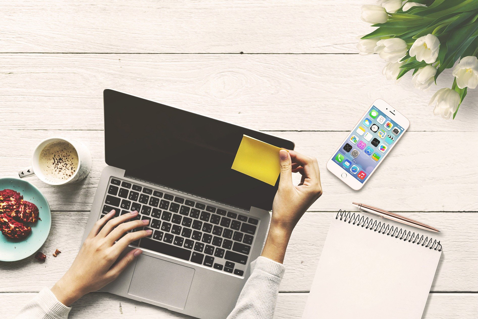 Obrazek przedstawia różne rzeczy leżące na stole. Są to kwiaty, pusta strona notesu, długopis, smartfon wyświetlający listę aplikacji, ciastka, kubek z kawą oraz laptop. Jedna ręka przykleja na ekran karteczkę, a druga leży na klawiaturze. Ekran jest wygaszony.