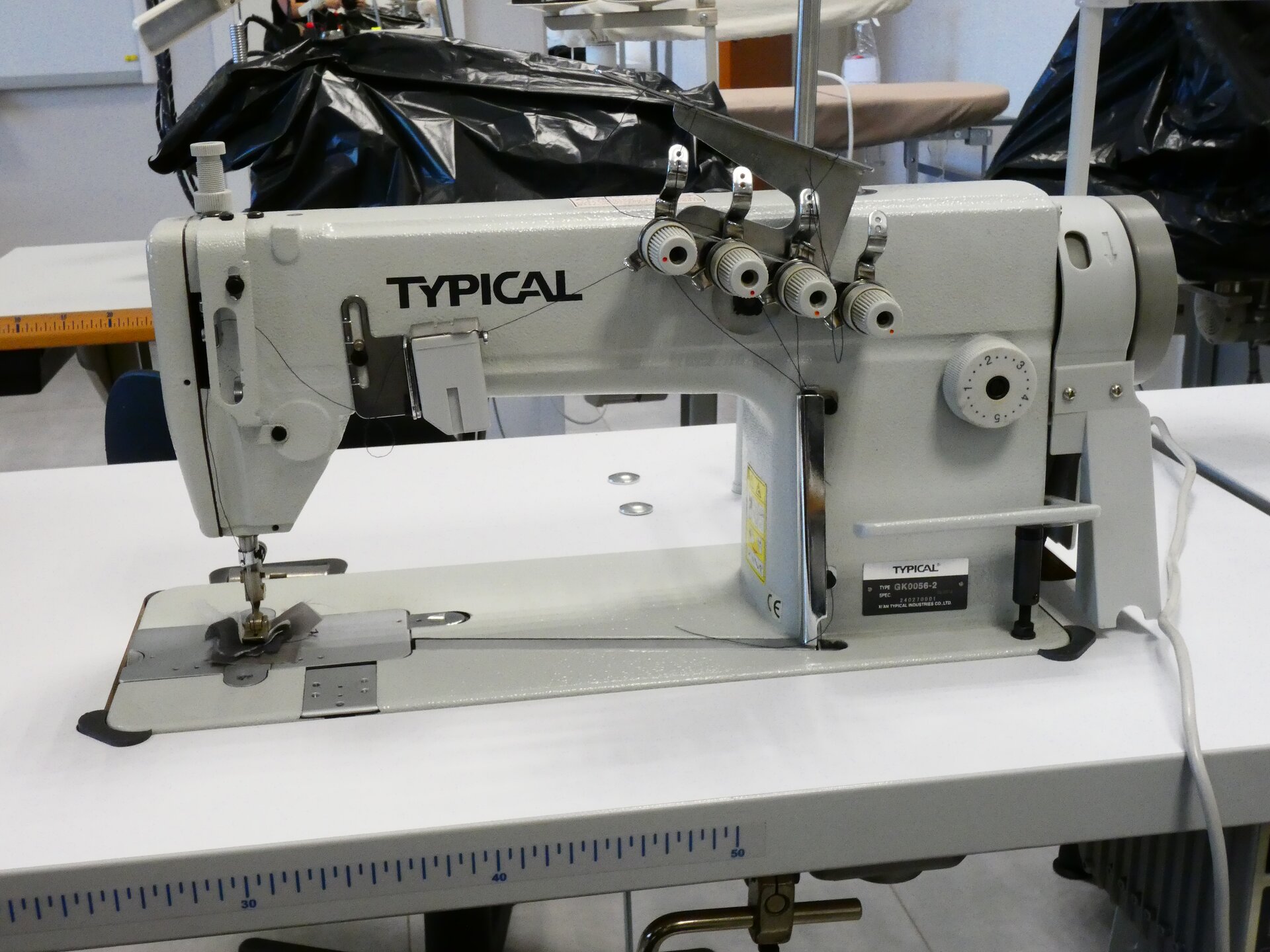 Zdjęcie przedstawia białą maszynę. Z lewej strony znajduje się ramię z igłą, z prawej z przodu pokrywy umieszczone zostały cztery małe pokrętła i jedno duże.