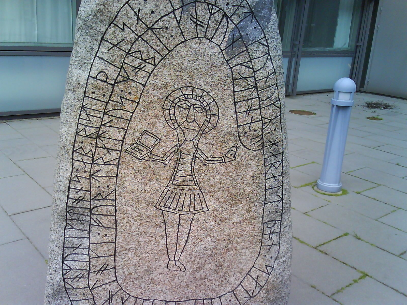 Zdjęcie współczesne, przedstawiające owalny kamień z dwoma wersami tekstu zapisanych runami po obwodzie kamienia. W centrum widoczna, zarysowana postać męska z aureolą na głowie, trzymająca odpowiednio w lewej i prawej dłoni telefon komórkowy i laptop. 