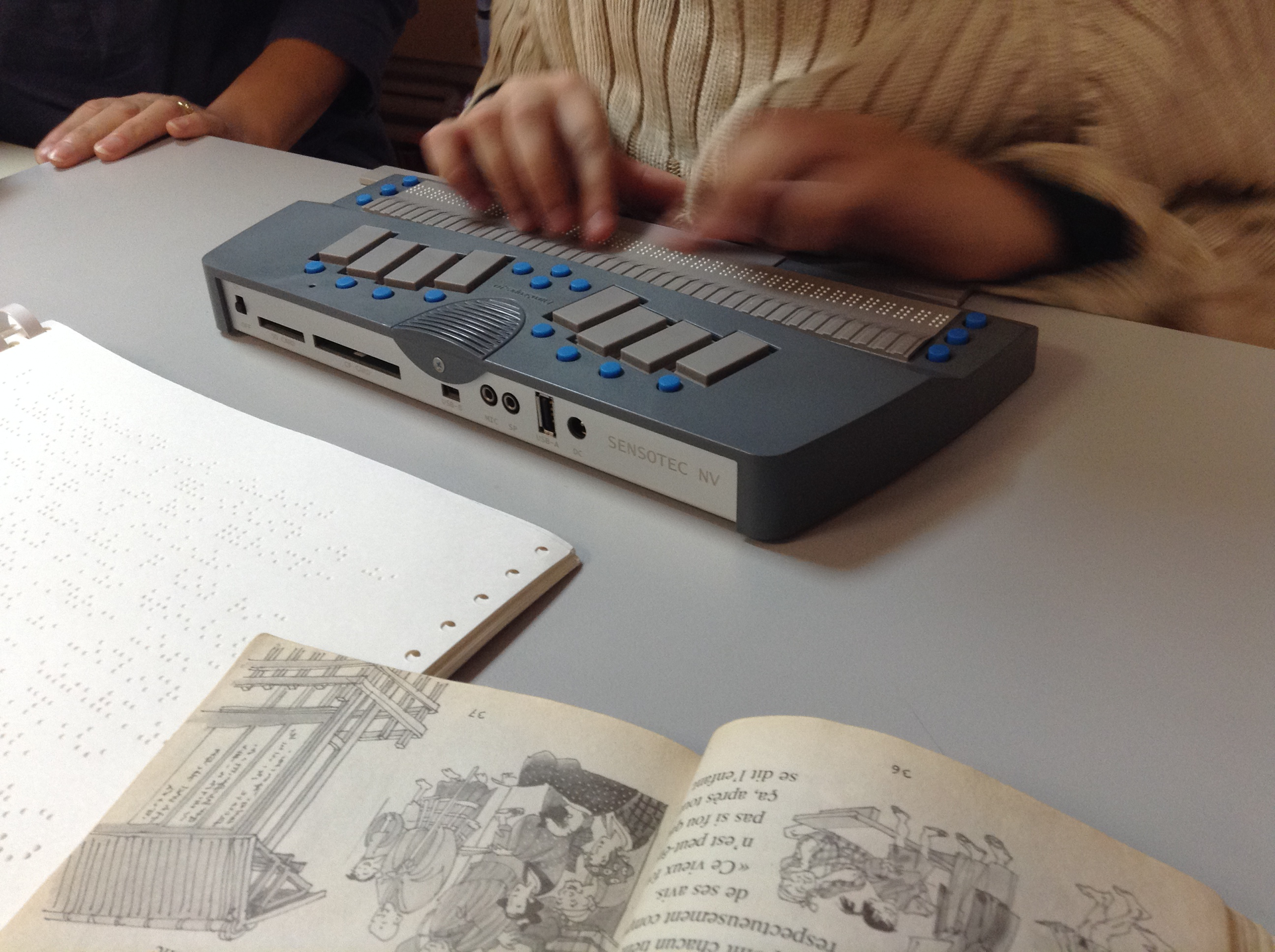 Na ilustracji znajduje się przyrząd o kształcie prostopadłościanu. Na górnej ścianie znajduje się rząd zapisany pismem Braille'a, nad każdą literą zapisaną brailem znajduje się prostokątny, czarny przycisk. Wyżej widać dwie grupy po cztery szare przyciski, ułożone tak, by łatwo było oprzeć o nie palce obydwóch rąk. Poniżej znajduje się kartka zapisana alfabetem Braille'a obok znajduje się książka z rysunkami.