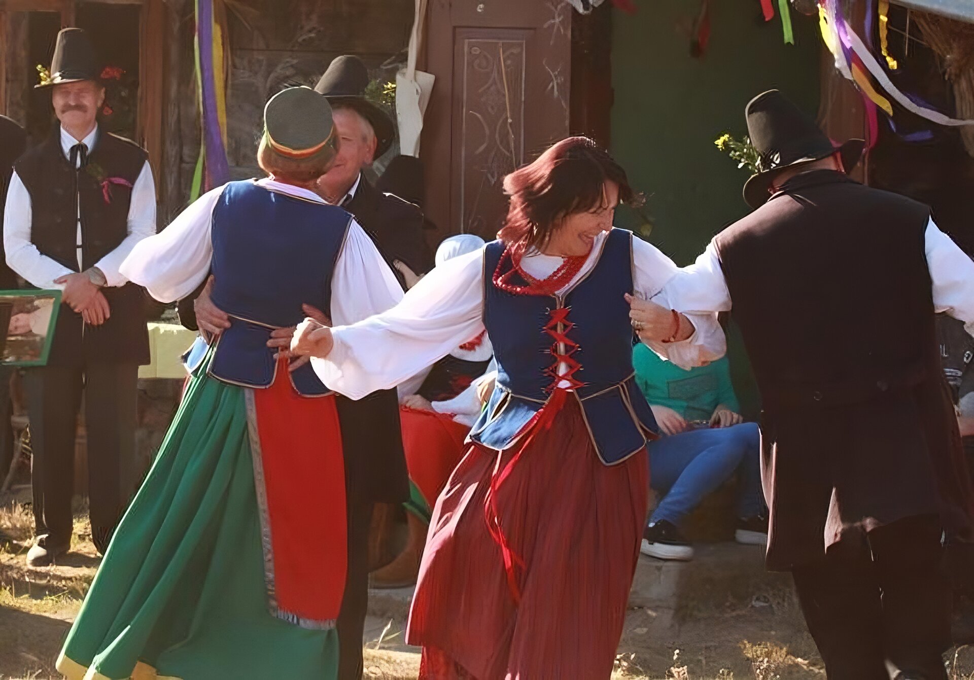 Zdjęcie przedstawia tańczących ludzi ubranych w stroje ludowe. Widoczne są dwie pary tańczące oraz jeden mężczyzna stojący po lewej stronie zdjęcia. Mężczyzna ubrany jest w czarne, długie spodnie, białą koszulę, czarną kamizelkę i czarny kapelusz. Pierwsza z tańczących par, znajdująca się bliżej stojącego mężczyzny, patrzy sobie w oczy. Kobieta stoi tyłem do fotografa. Ubrana jest w zieloną spódnicę, czerwony fartuch, białą koszulę i niebieską kamizelkę. Na głowie ma szary kapelusz. Kobieta niemal całkowicie zasłania partnera - widoczna jest tylko jego czarna marynarka oraz czarny kapelusz. Po prawej stronie widoczna jest druga para, która trzyma się za łokcie. Kobieta ma czerwoną spódnicę, białą koszulę, niebieską kamizelkę oraz czerwone korale. Ma rude, rozpuszczone włosy. Śmieje się. Mężczyzna jest odwrócony do obiektywu tyłem. Ma czarne spodnie, białą koszulę, czarną kamizelkę i czarny kapelusz, do którego przypiął żółty kwiat. W tle widoczny jest fragment budynków. Ludzie stoją na krótkiej, zielonej trawie. Zdjęcie zostało zrobione w świetle dziennym.