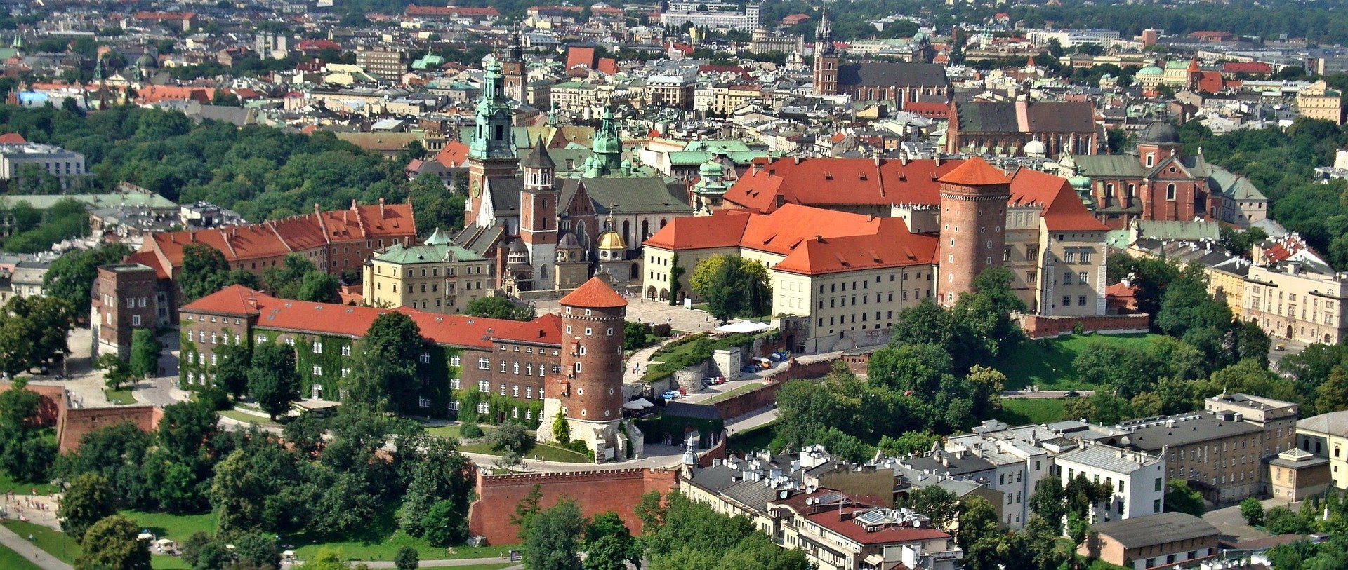 Ilustracja przedstawia zdjęcie Zamku Królewskiego i Wawelu w Krakowie z lotu ptaka. Widoczne charakterystyczne mury, wieże oraz budynki najstarszej części Krakowa widnieją nad miastem. W oddali widać panoramę Krakowa. 