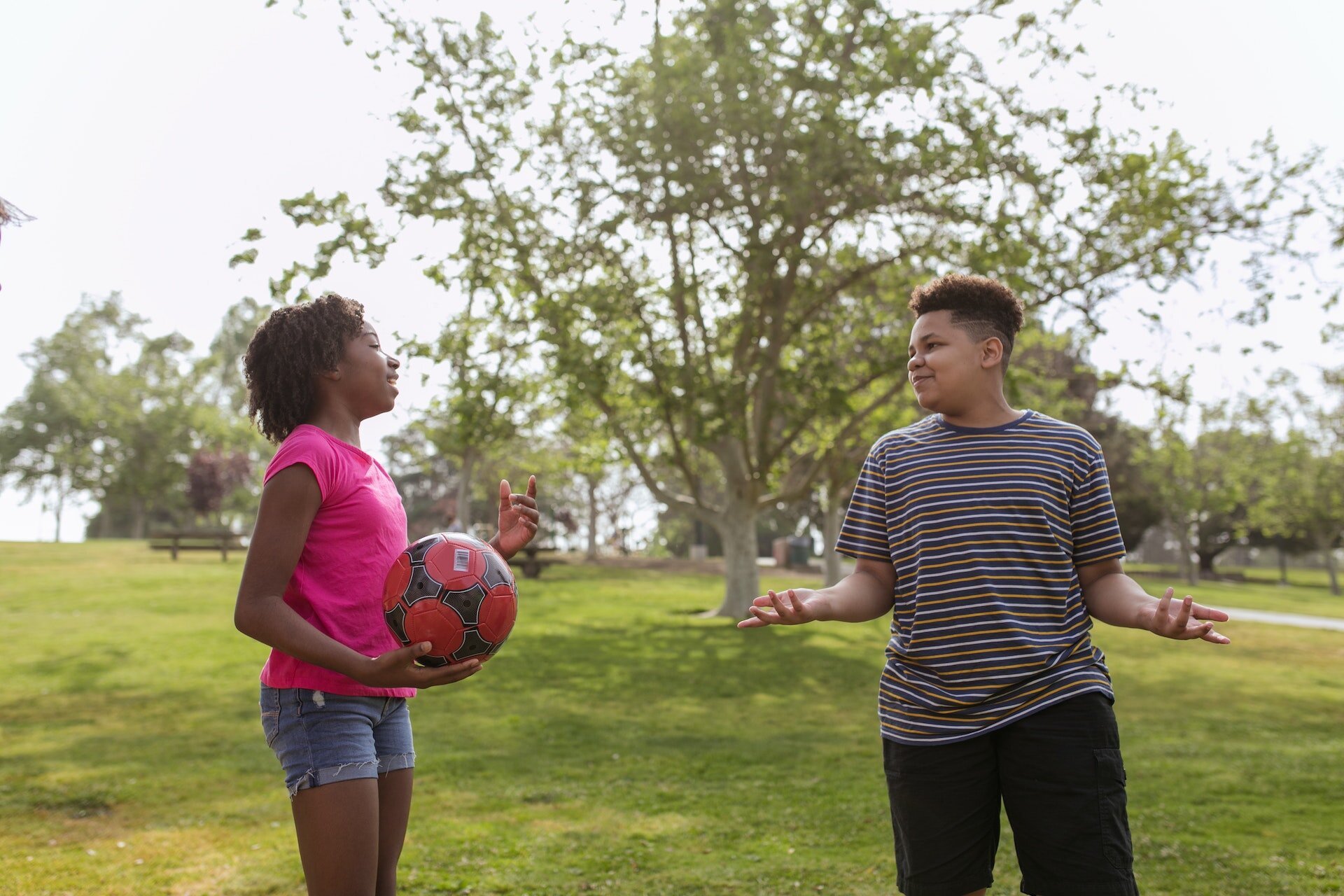 Zdjęcie przedstawia dwie młode osoby: dziewczynę ubraną w bluzkę z krótkim rękawem i szorty, która trzyma przed sobą piłkę, oraz chłopaka w pasiastej koszulce i dresowych spodniach. Dziewczyna coś mówi, a chłopak rozkłada ręce.