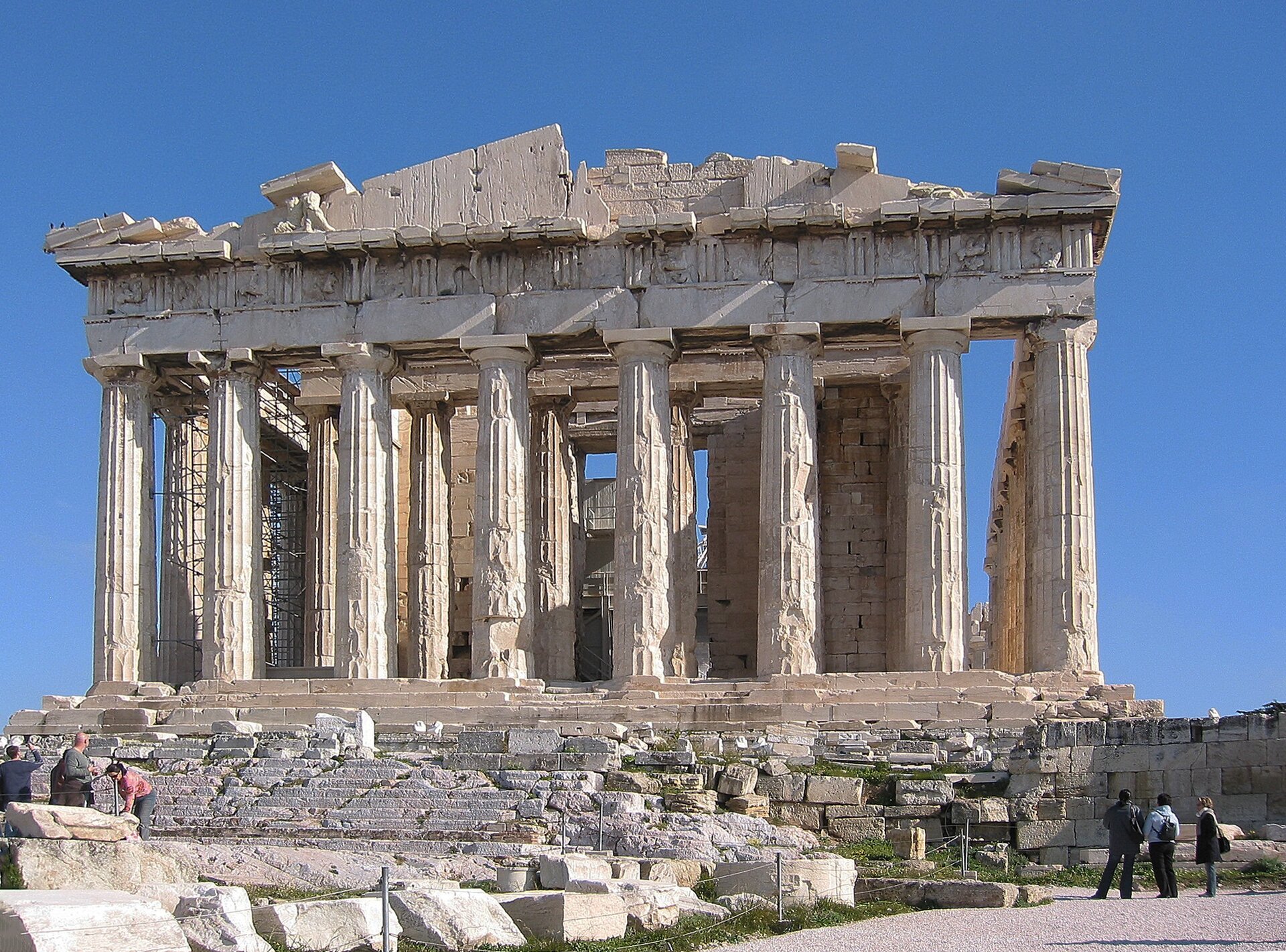Fotografia nieznanego autora przedstawia widok na Partenon – świątynię poświęconą bogini Atenie. Budynek został zbudowany z jasnych cegieł i wsparty na kolumnadzie. Budynek jest zniszczony i nadgryziony zębem czasu. Obok budynku widoczni są turyści. Fotografia została wykonana w jasny i bezchmurny dzień. 