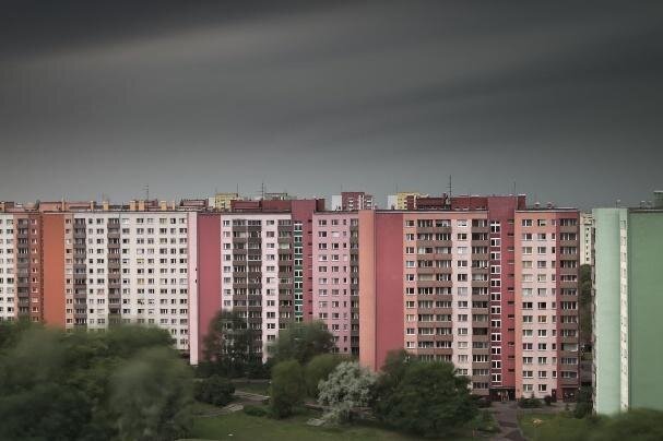 Zdjęcie przedstawia kilka wysokich bloków. Każdy z nich pomalowany jest w odcieniach różu. Przed nimi rosną drzewa. 