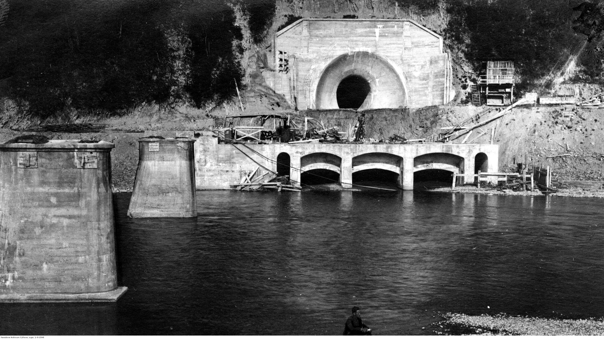 Zdjęcie przedstawia betonową budowlę w wodzie. Wydrążona jest w niej dziura. Pod nią w betonie są trzy łuki. Naokoło znajdują się rusztowania oraz rozkopana jest ziemia. Ponad wodą z lewej strony uwidocznione są dwie betonowe bryły. Teren otaczają rośliny. Ilustracja jest czarno‑biała.