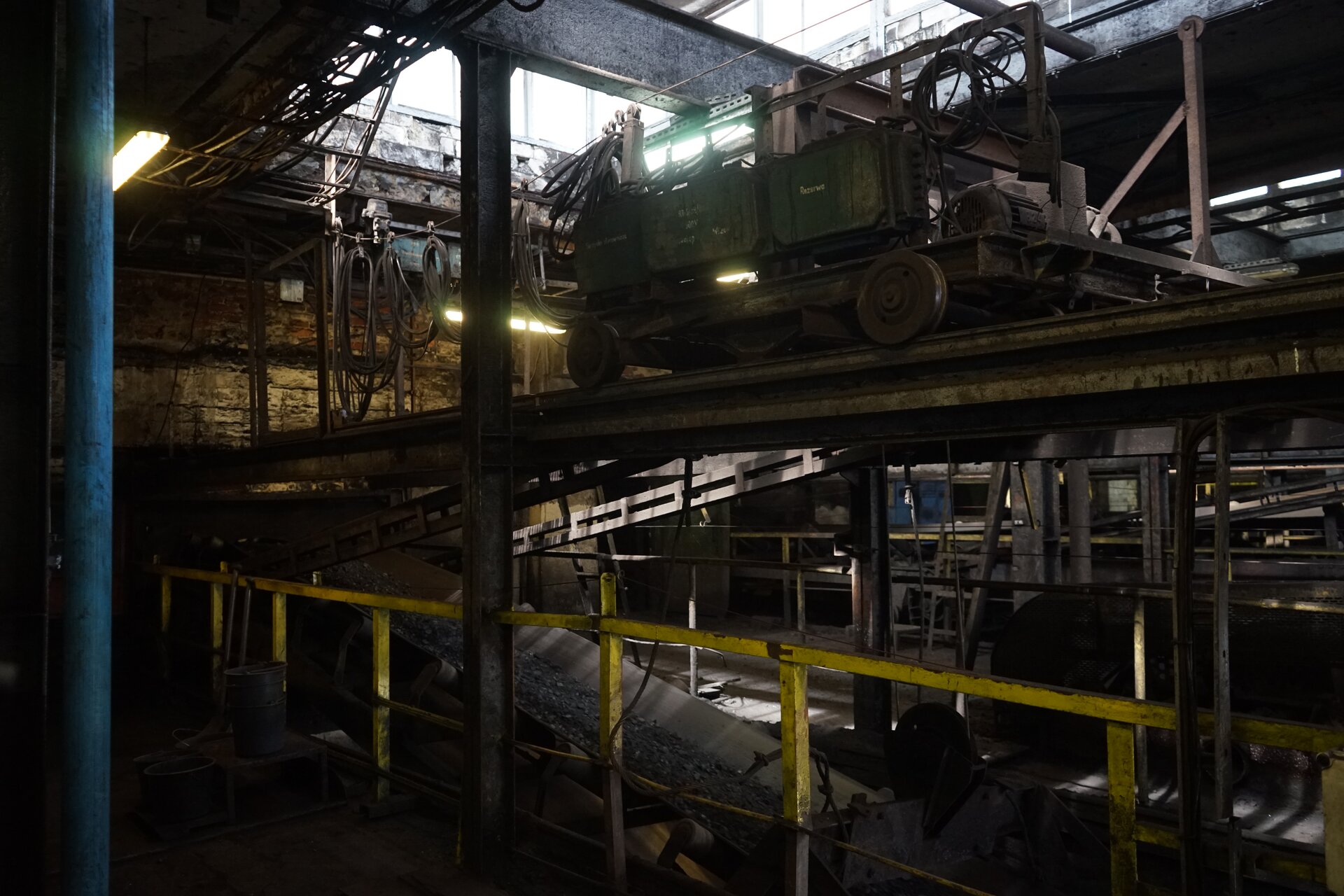 Na zdjęciu widoczna jest ładowarka węgla na wagony. W dolnej części zdjęcia widoczny jest przenośnik taśmowy. Na taśmie przenośnika transportowany jest węgiel. Nad przenośnikiem widoczne są tory. Tory są osadzone na stalowej rampie. Na torach stoi skład. Skład jest częściowo zdemontowany. Z przodu składu widać jednostkę napędową. Napęd jest o kształcie cylindra na powierzchni którego widać wycięcia o kształcie harmonijki. W tle zdjęcia widać kable. Poniżej zdjęcia widoczny jest przycisk multimedialny z nagraniem audio. Treść nagrania jest zgodna z tytułem zdjęcia.