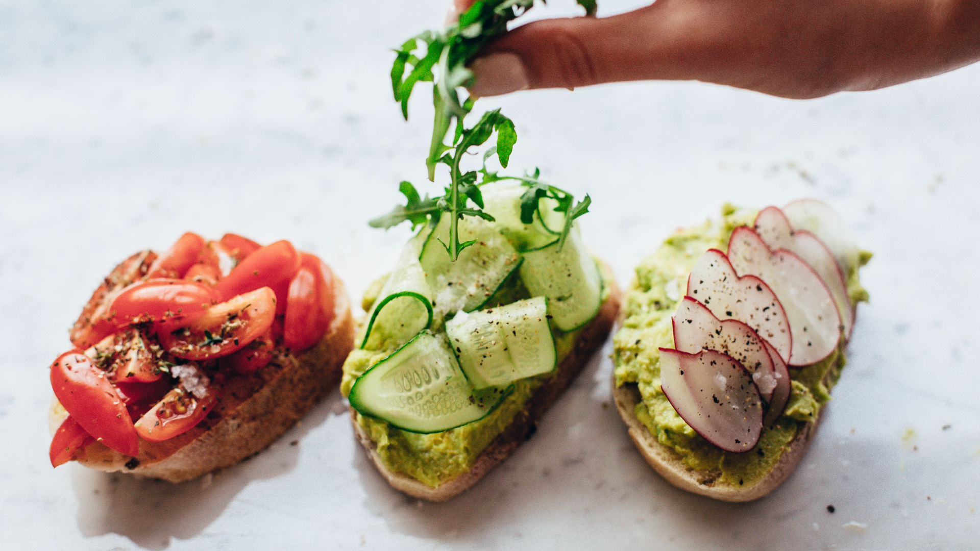 Zdjęcie przedstawia trzy przygotowane kanapki. Wszystkie kanapki są z jakąś pastą. Na pierwszej znajdują się pomidory, na drugiej plasterki ogórka, a na trzeciej plasterki rzodkiewki.