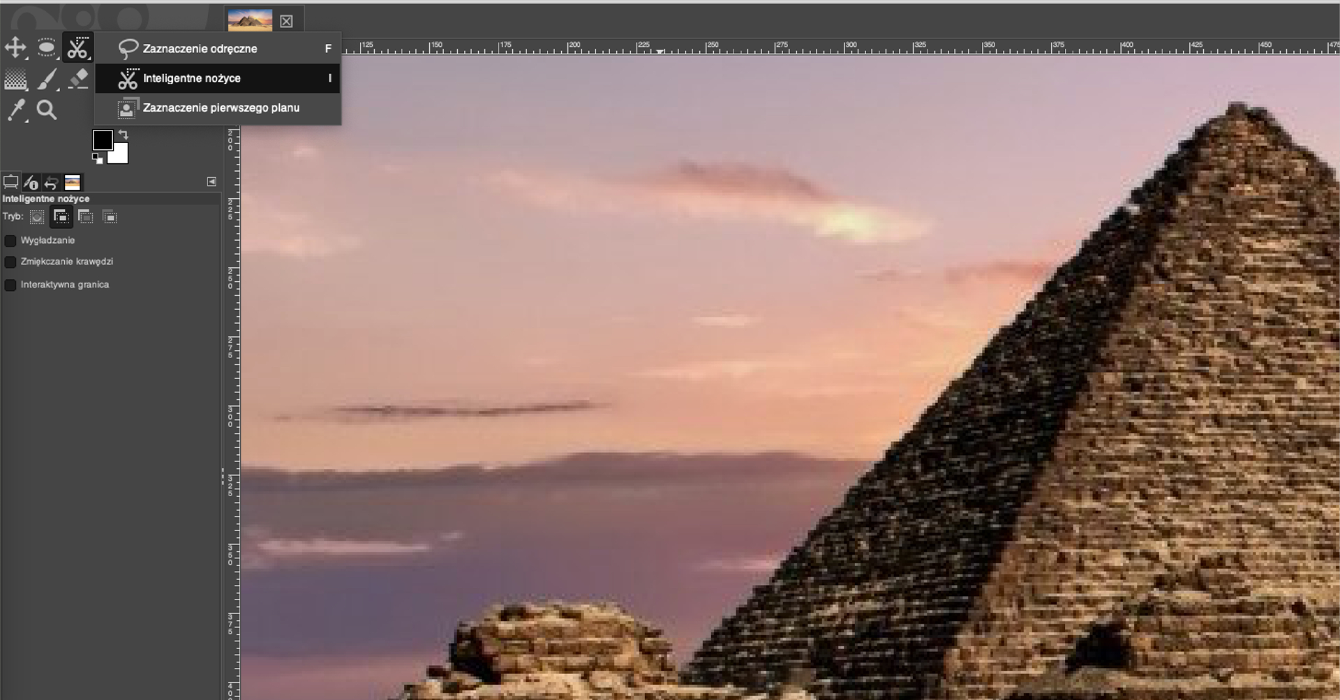 Ilustracja przedstawia okno w programie GIMP. Wewnątrz okna znajduje się fragment zdjęcia z piramidą – wierzchołek najwyższej piramidy oraz fragment nieba. Po lewej stronie znajduje się pasek z menu programu. Wybrana jest miniatura przedstawiająca nożyczki i rozwinięta lista z pozycjami: Zaznaczenie odręczne, Inteligentne nożyce, Zaznaczenie pierwszego planu. Wybrana jest opcja: Inteligentne nożyce. Całe zdjęcie piramidy jest otoczone miarką z zaznaczonymi wartościami liczbowymi.