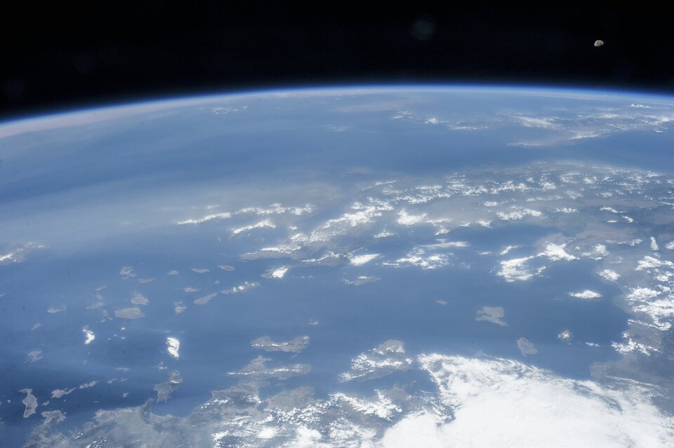 Zdjęcie przedstawia fragment powierzchni Ziemi sfotografowany z orbity. W centralnej i dolnej części kadru obszar mórz poprzecinanych skrawkami lądu, w części górnej zakrzywiona linia horyzontu z niebieskim paskiem atmosfery nad którą rozpościera się czerń kosmosu. W prawym górnym rogu kadru mały Księżyc.