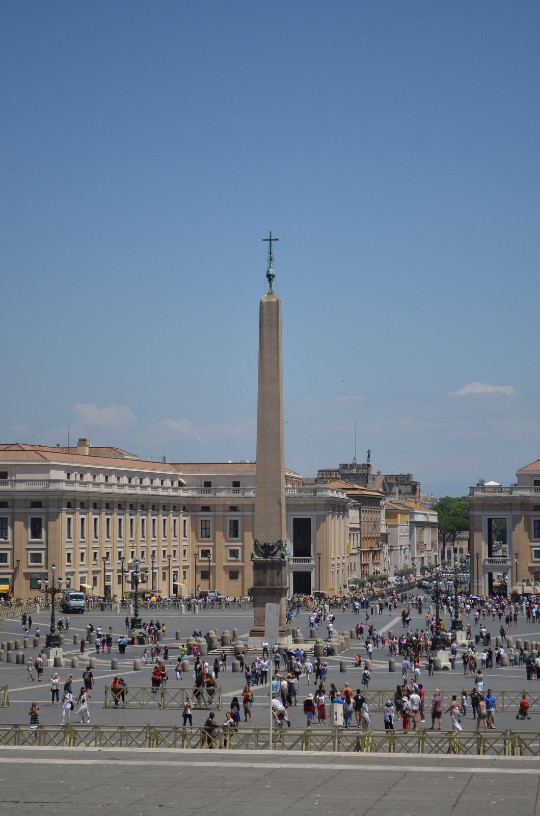 Ilustracja przedstawia obelisk na Placu świętego Piotra w Watykanie. Pośrodku placu, na którym chodzą ludzie, znajduje się obelisk zwieńczony krzyżem. W tle widoczna jest architektura.