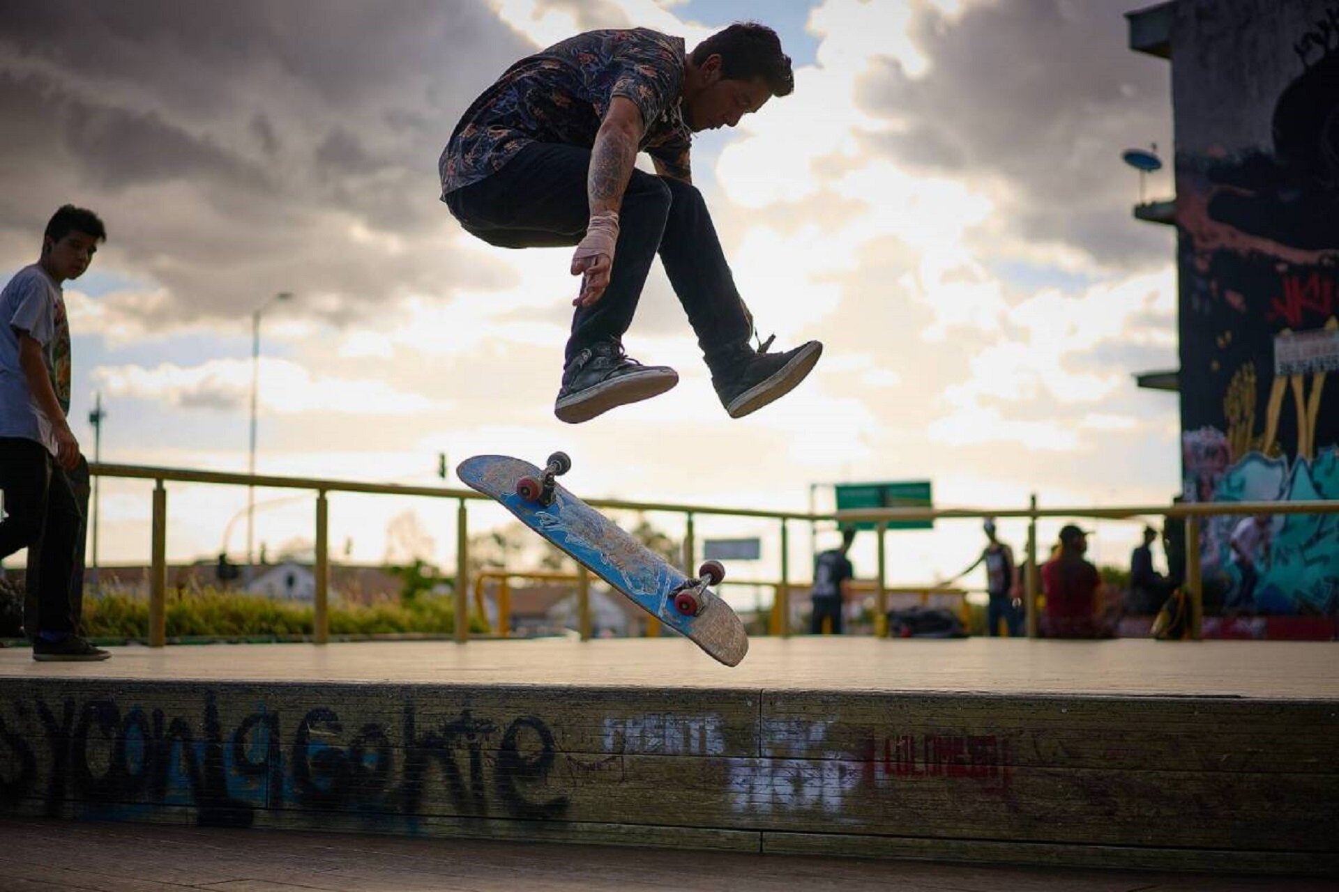 Ilustracja przedstawia skatera, młodego mężczyznę wykonującego trik na deskorolce. W tle widoczny skatepark.