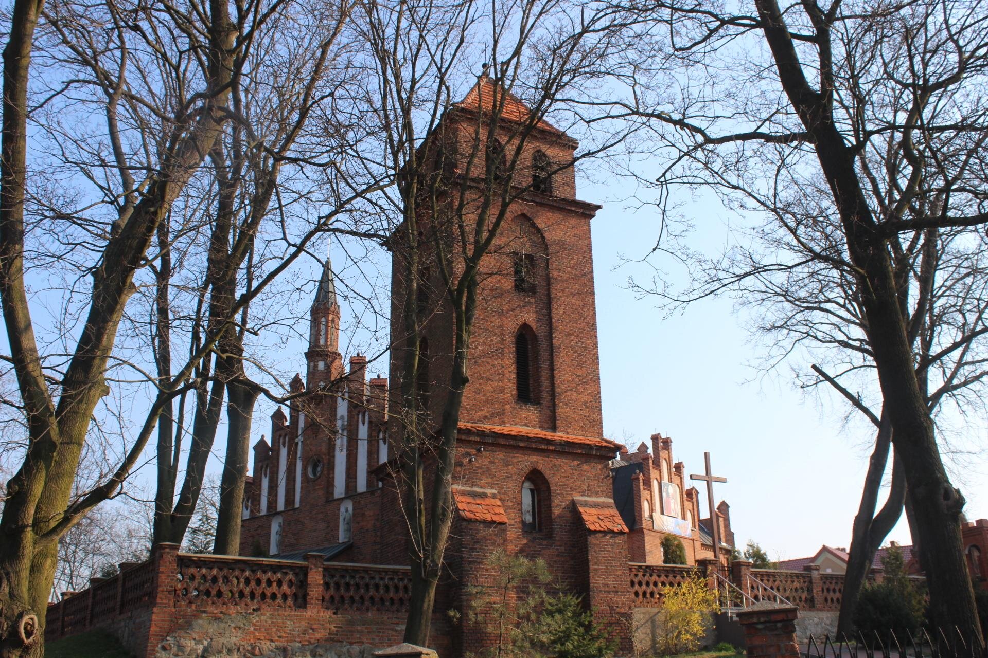 Zdjęcie przedstawia wysoką, wykonaną z czerwonej cegły, wieżę. Na każdym z trzech pięter znajduje się jedno okno, a na jej szczycie, na piętrze czwartym, dwa okna. Z obu stron wieży mur, a w tle fragmenty kościoła. Wokół wieży drzewa bez liści.