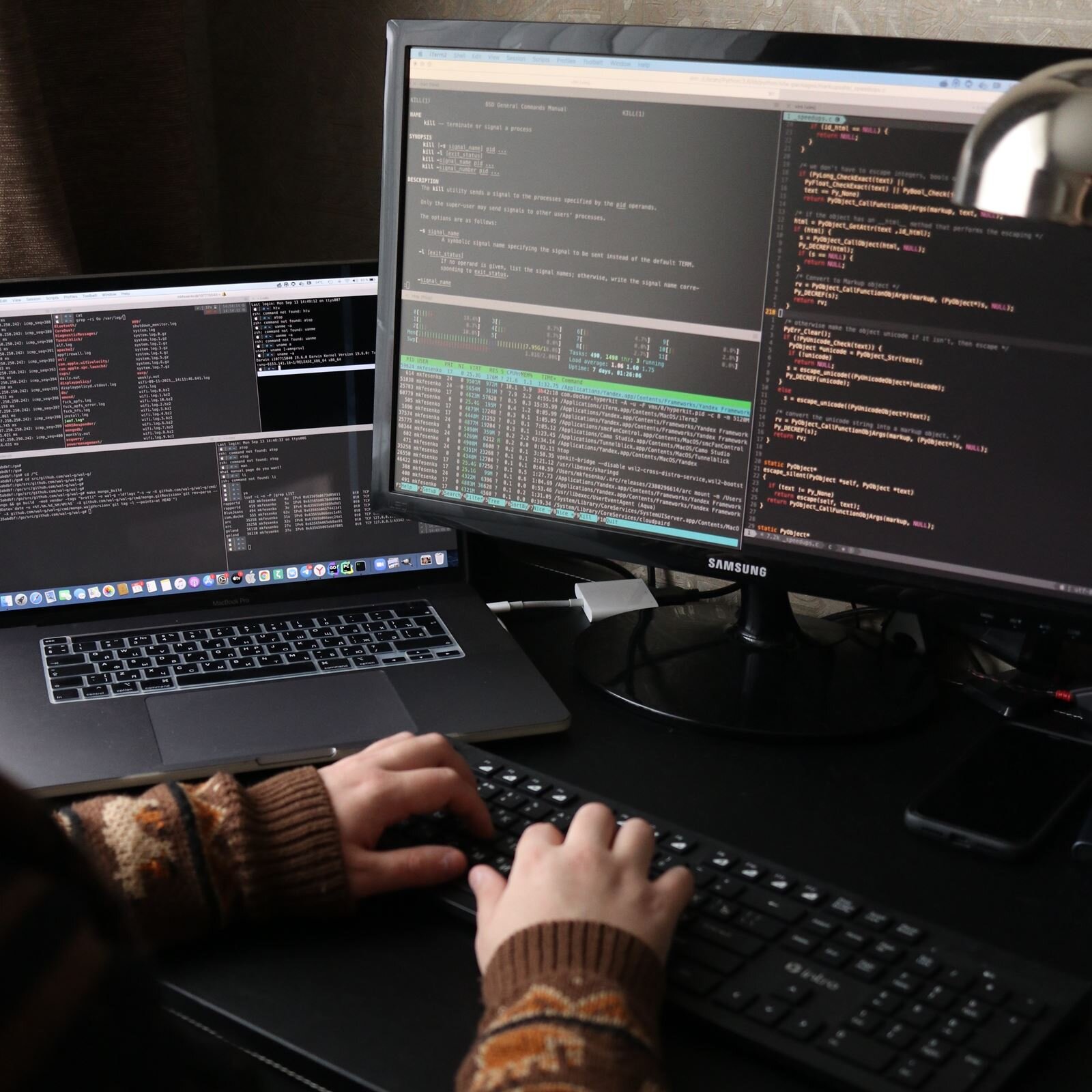 Zdjęcie przedstawia biurko, na którym po prawej stronie widoczny jest monitor komputera stacjonarnego, zaś po lewej stronie widoczny jest otwarty laptop. Przed monitorem komputera stacjonarnego widoczna jest klawiatura, na której spoczywają dłonie. Na monitorach obydwu komputerów widoczne są różnego rodzaju kody, otwarte foldery, zapisy.