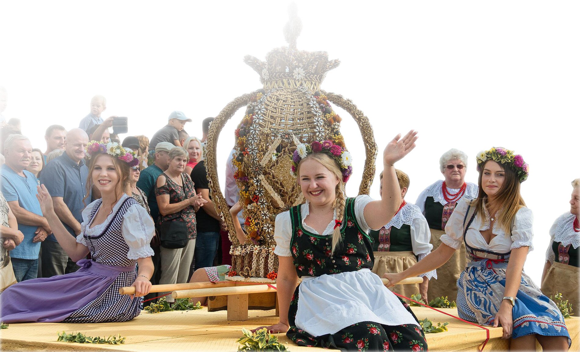 Zdjęcie przedstawia moment przykładowego wydarzenie z elementami lokalnego folkloru. Na zdjęciu widać kobiety ubrane w stroje ludowe siedzące na drewnianej platformie oraz ludzi obserwujących paradę. Pośrodku platformy stoi słomiana figura.