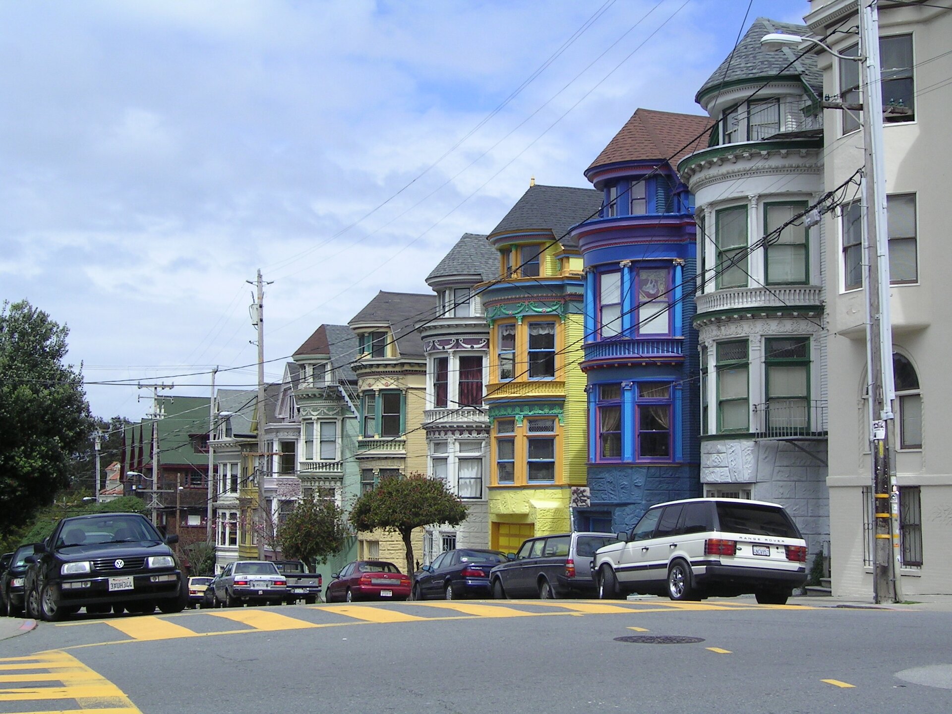 Szeroka ulica z pasami, wzdłuż niej szereg kolorowych budynków o zaokrąglonych kształtach, mnóstwo okien. Na ulicy jadące samochody.
