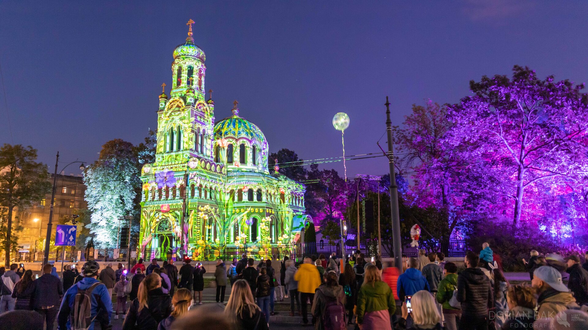 Na zdjęciu znajduje się budynek z wieżą i kopułą i wyświetlane są na nim kolorowe wzory przedstawiające trawę i kwiaty. Po lewej stronie stoi wysokie drzewo liściaste podświetlone na zielono. Po prawej stronie rośnie kilka drzew liściastych podświetlonych na fioletowo oraz świecący, biały balon. Przed budynkiem stoi tłum ludzi. Tło zdjęcia stanowi zabudowa miejska i ciemne niebo.