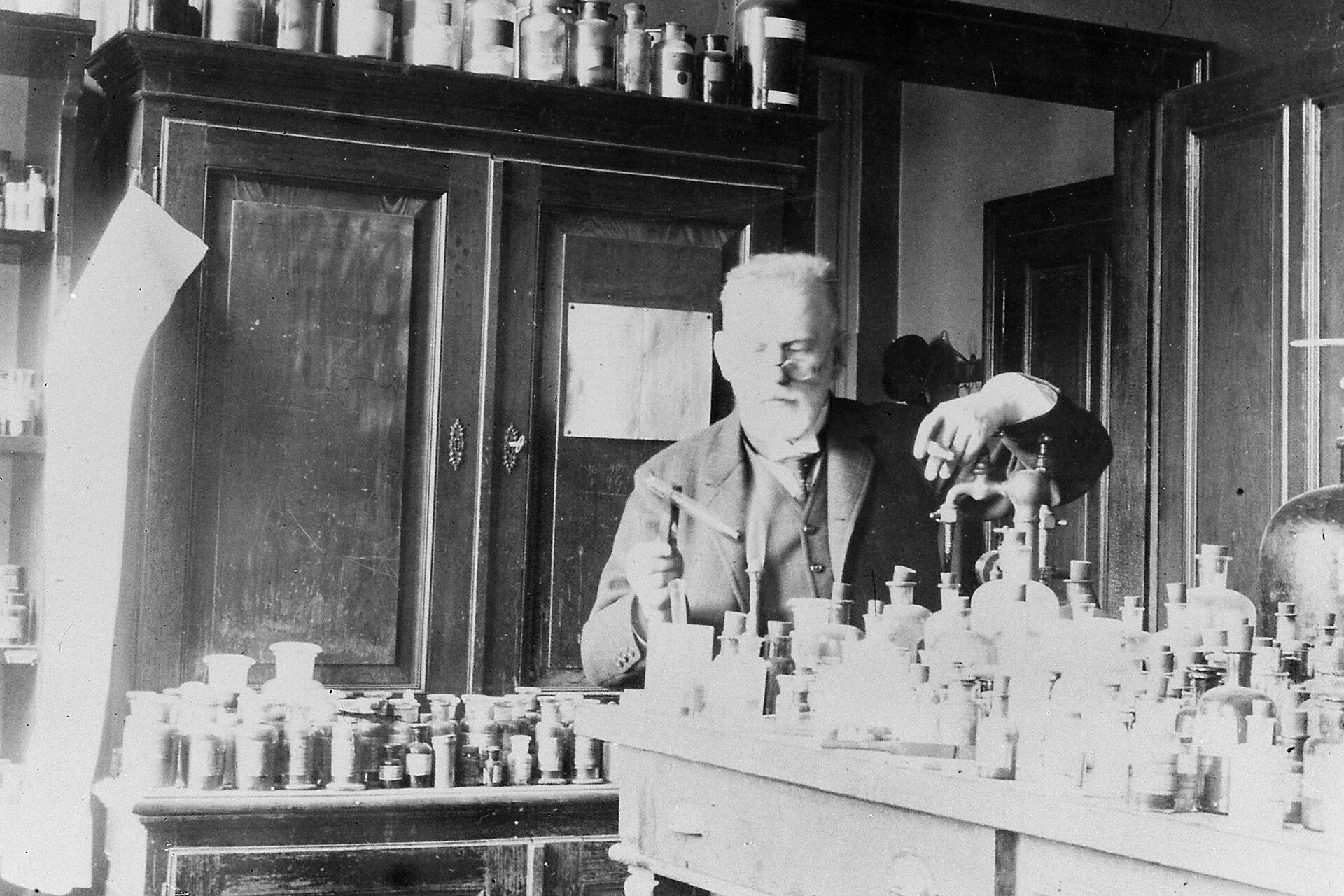 Zdjęcie czarno-białe. Za stołem, zapełnionym szklanymi butelkami różnej wielkości, stoi Paul Ehrlich - niemiecki chemik i bakteriolog. Ten siwy, brodaty mężczyzna ubrany jest w ciemną marynarkę z kamizelką. Na czubku nosa okrągłe, druciane okulary. Pod sztywnym, lekko uniesionym kołnierzem białej koszuli przewiązany krawat. Uczony w swej prawej dłoni  w szczypcach trzyma probówkę. Przygląda się jej z uwagą. Swoją lewą dłoń, trzymajacą cygaro, wspiera na kurku kranu. Kran cześciowo zasłonięty zakorkowanymi butelkami z białymi etykietami.  Za uczonym stoi kredens z porysowanymi drzwiczkami. Na jego półce i szczycie - rzędy zakorkowanych butelek z chemikaliami.