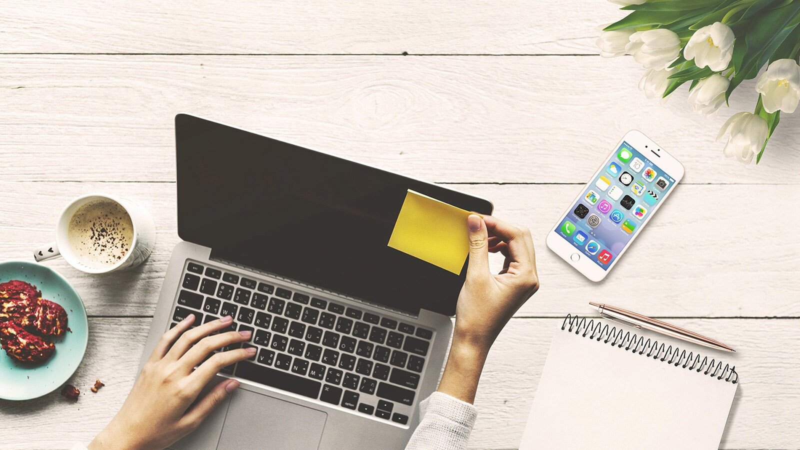 Zdjęcie przedstawia biurko, na którym widoczny jest laptop, do którego ekranu ktoś przylepia żółtą karteczkę. Obok leży telefon i stoi filiżanka z kawą oraz ciastka na talerzyku. 